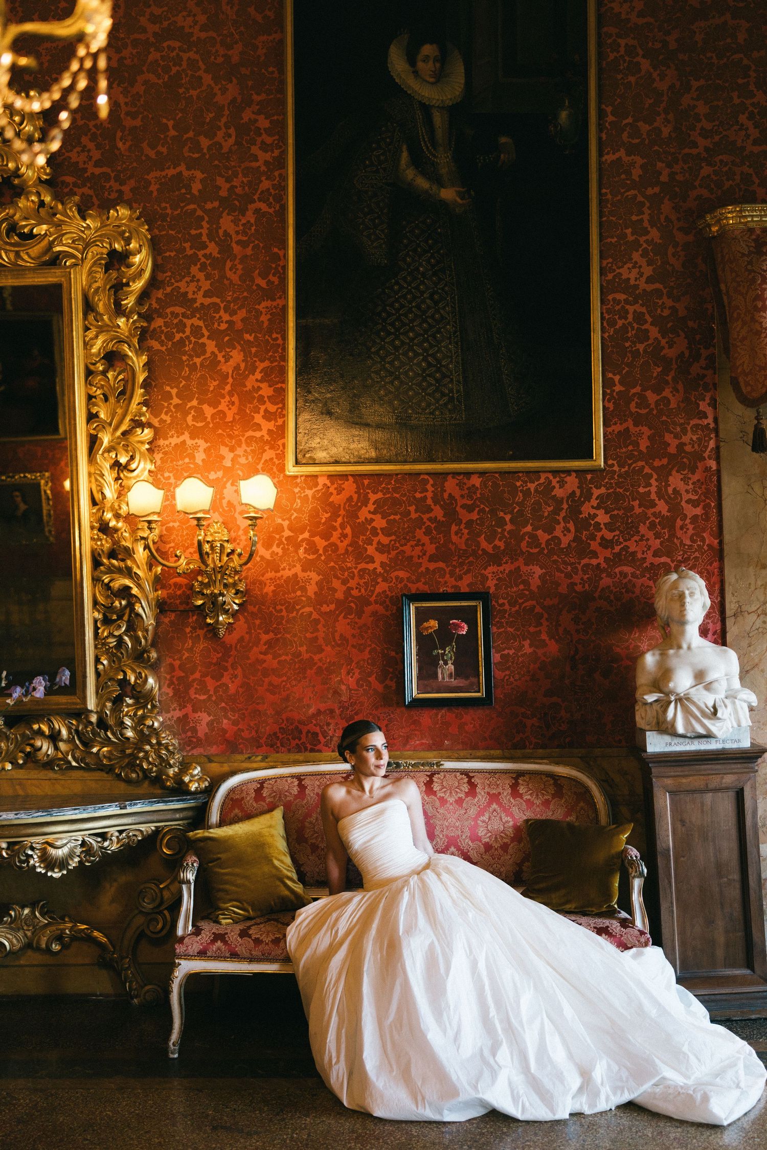 Bride in a white ball gown sitting on an ornate sofa in a grand room with red damask walls and gilded decor.
