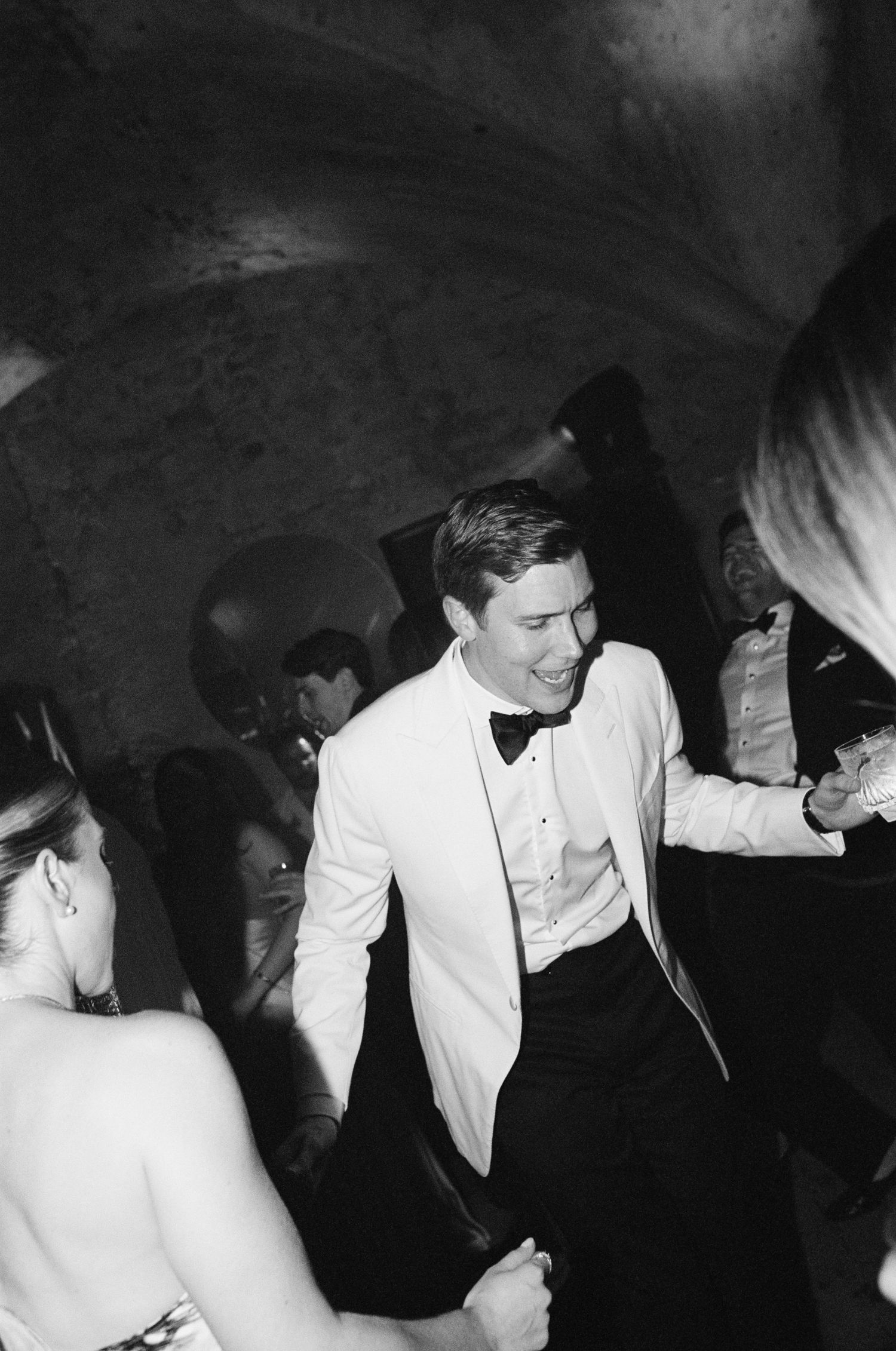 Groom in white tuxedo jacket dancing joyfully at wedding reception in stone venue.
