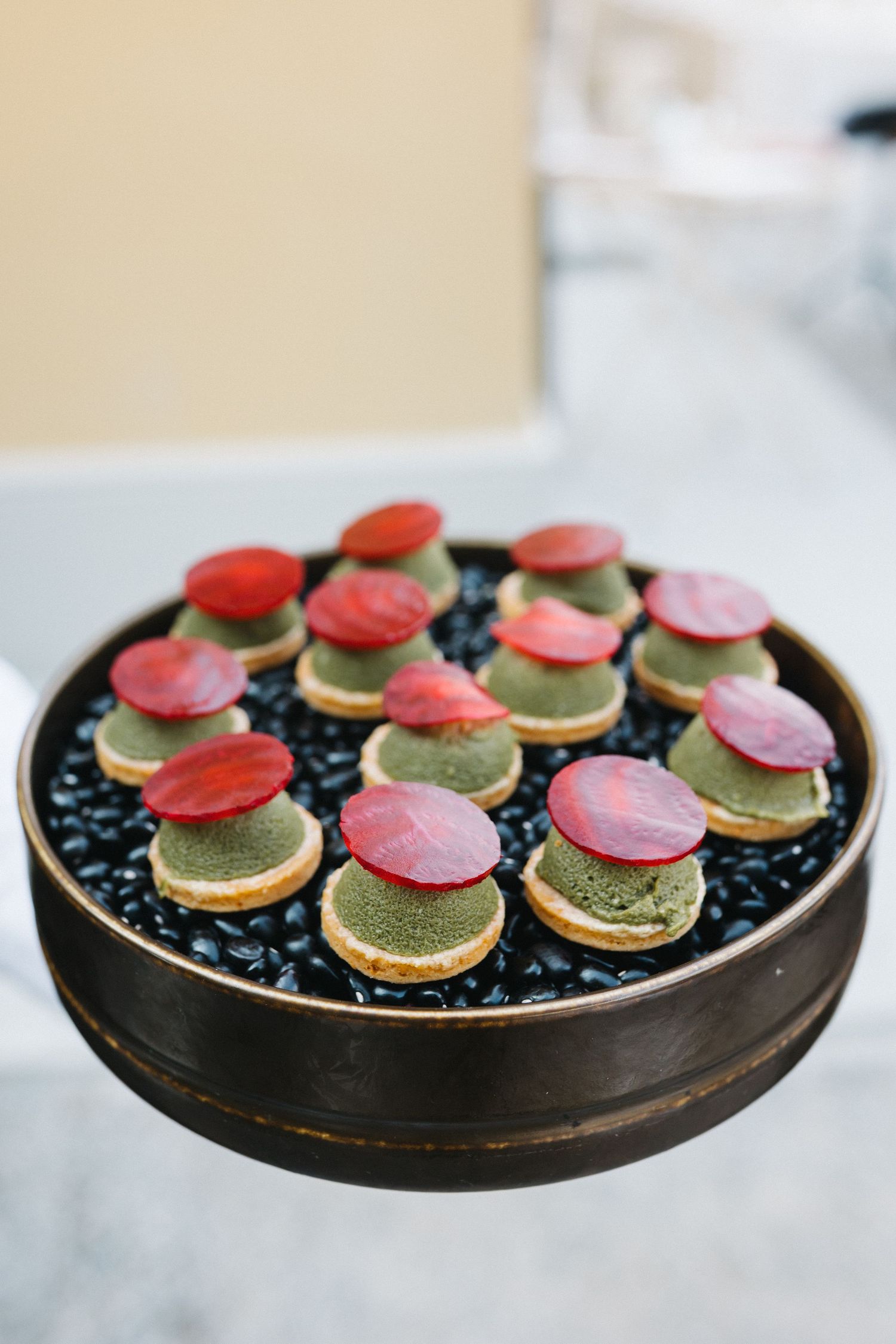 Elegant tray of matcha petit fours topped with red fruit slices, displayed on black beans at a catered event.