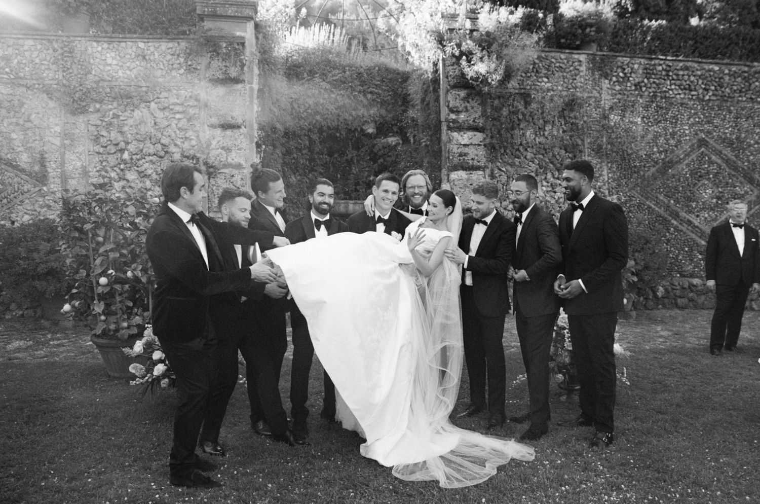 Groom carried by groomsmen in tuxedos during joyful outdoor wedding celebration in black and white.