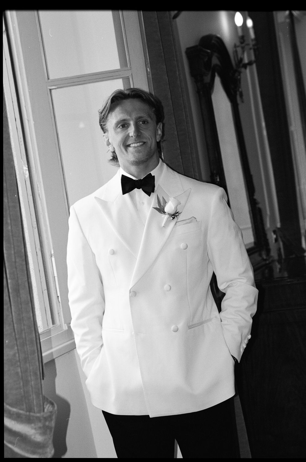 Elegant groom in white double-breasted tuxedo jacket with black bow tie smiling by a window in black and white.