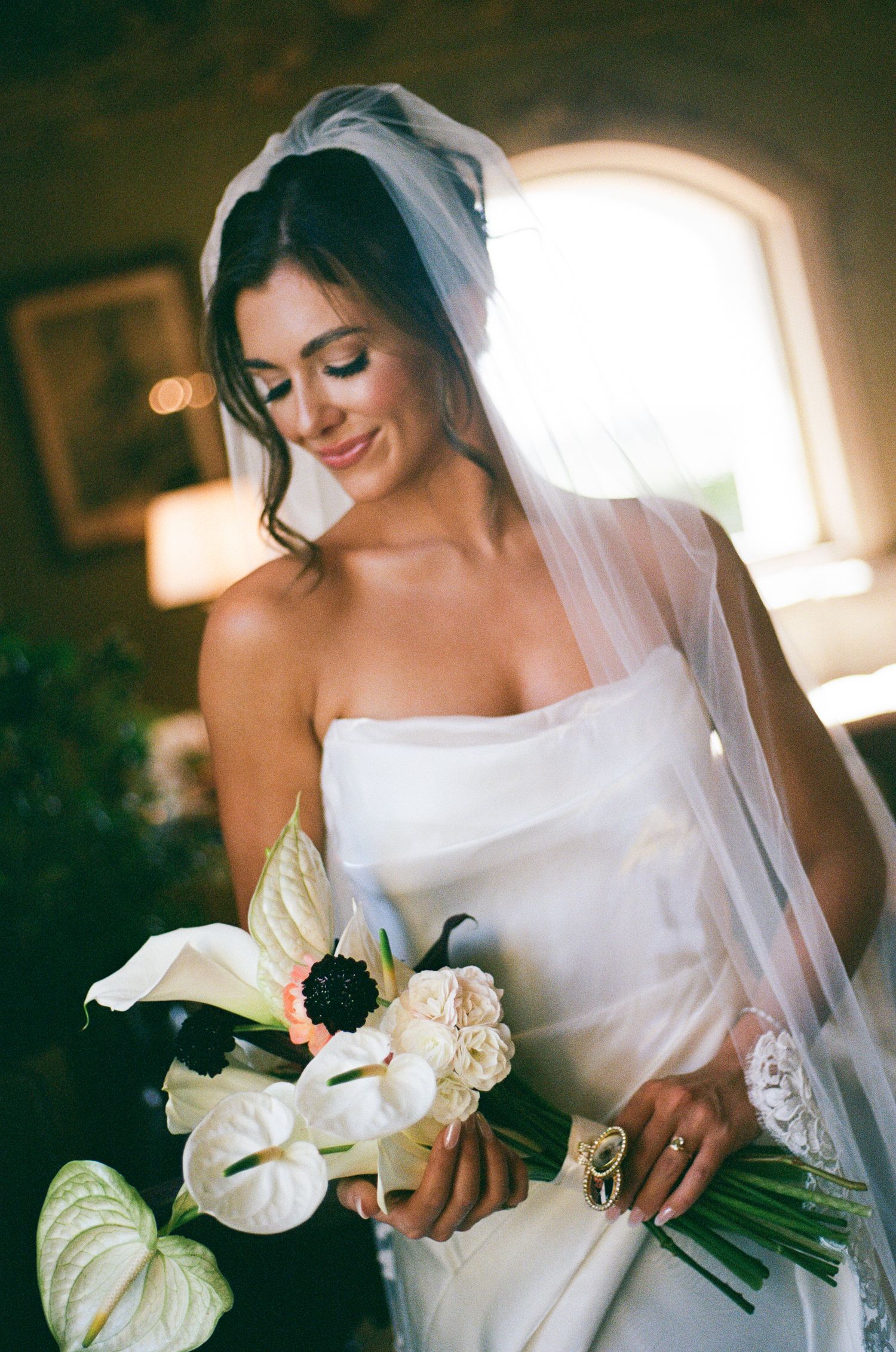 Radiant bride in strapless white gown holding a bouquet of white anthurium and roses by a window.