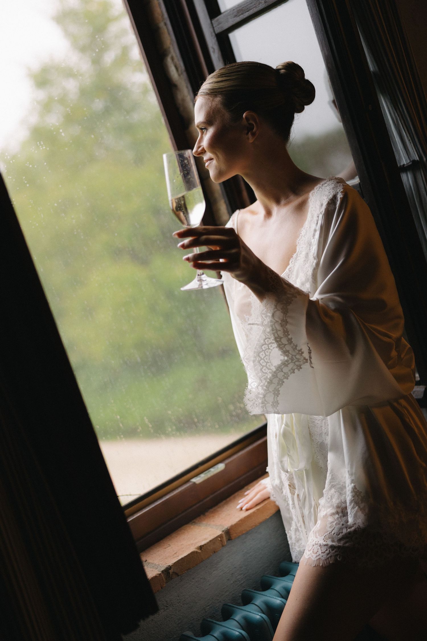 Bride in lace robe holding champagne glass gazing out rainy window on wedding day.