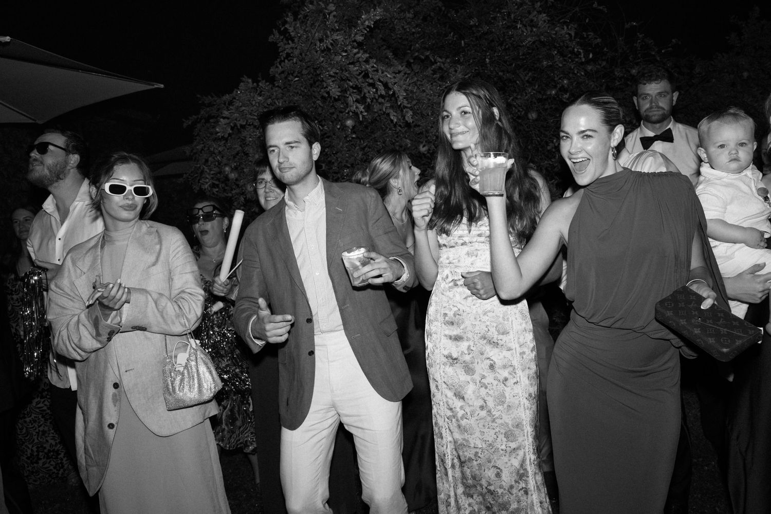 Black and white photo of elegantly dressed guests socializing at an outdoor evening party or social event.