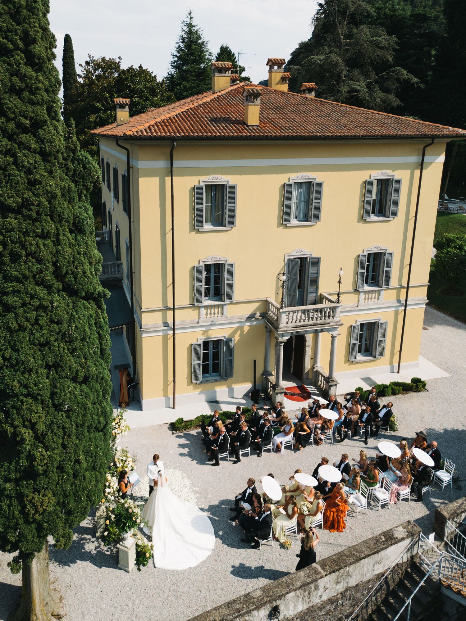 Aerial view of a wedding ceremony at a stunning yellow Italian villa with terracotta roof and lush cypress trees.