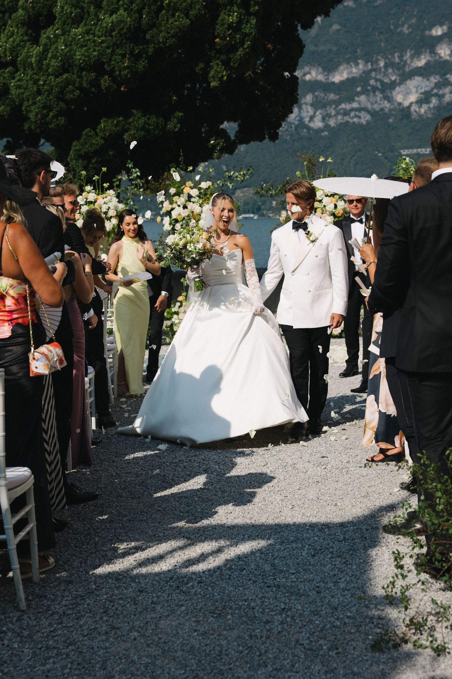 Bride and groom walk down outdoor aisle at Lake Como wedding ceremony, surrounded by guests and floral arrangements.