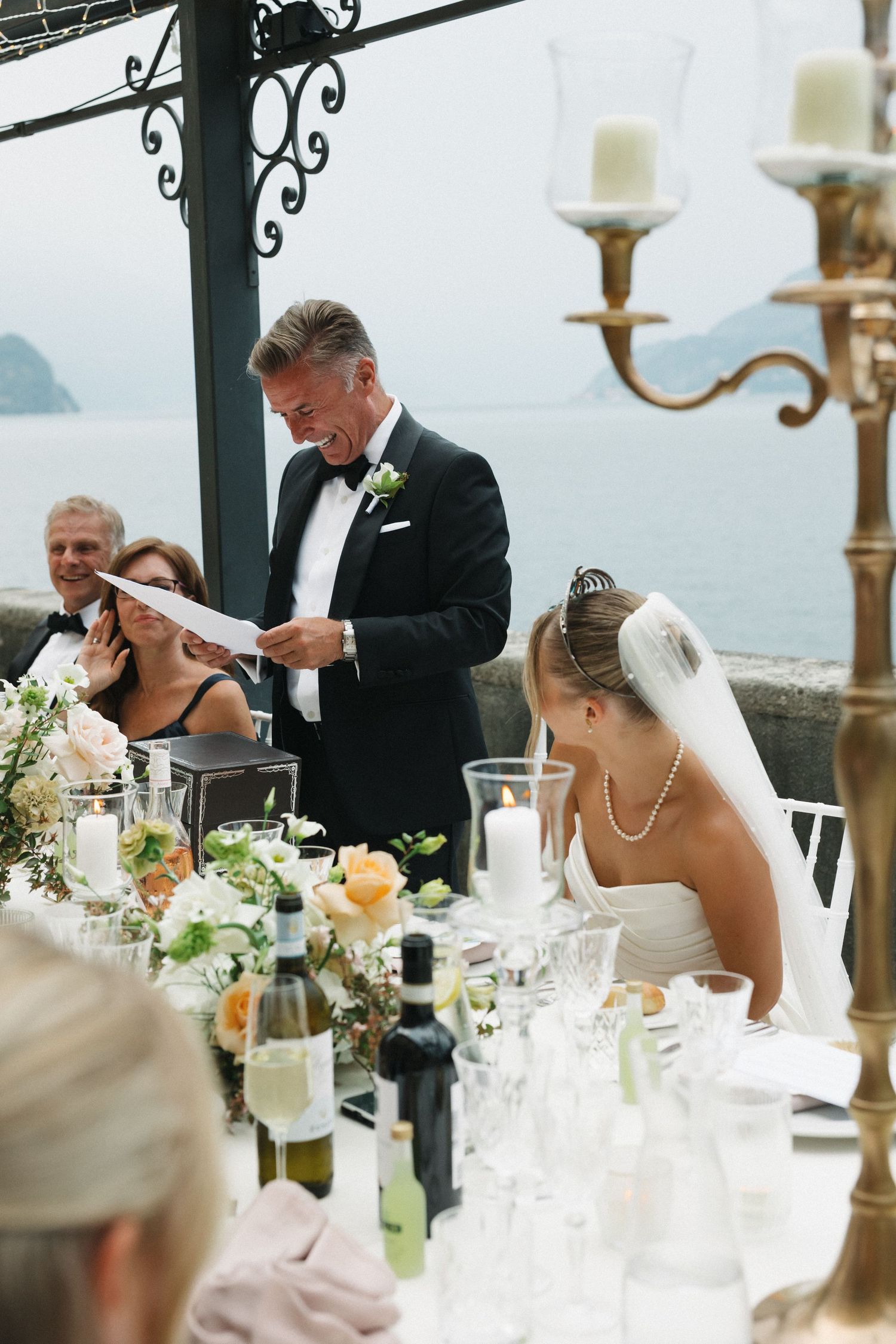 Father of the bride reads heartfelt speech at elegant outdoor lakeside wedding reception with bride in veil seated nearby.