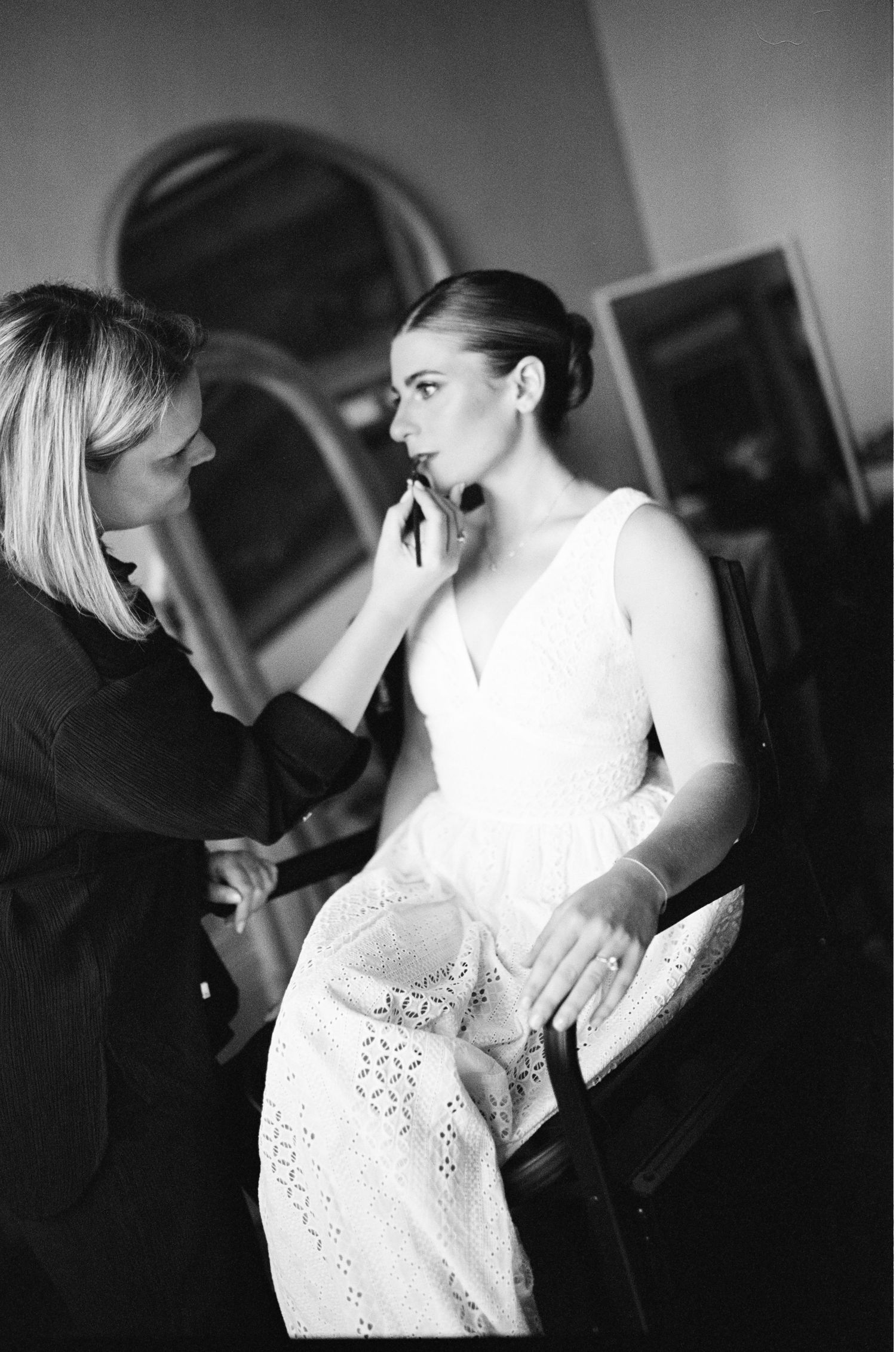 Makeup artist applies lipstick to bride in white dress seated in chair, black and white photo.