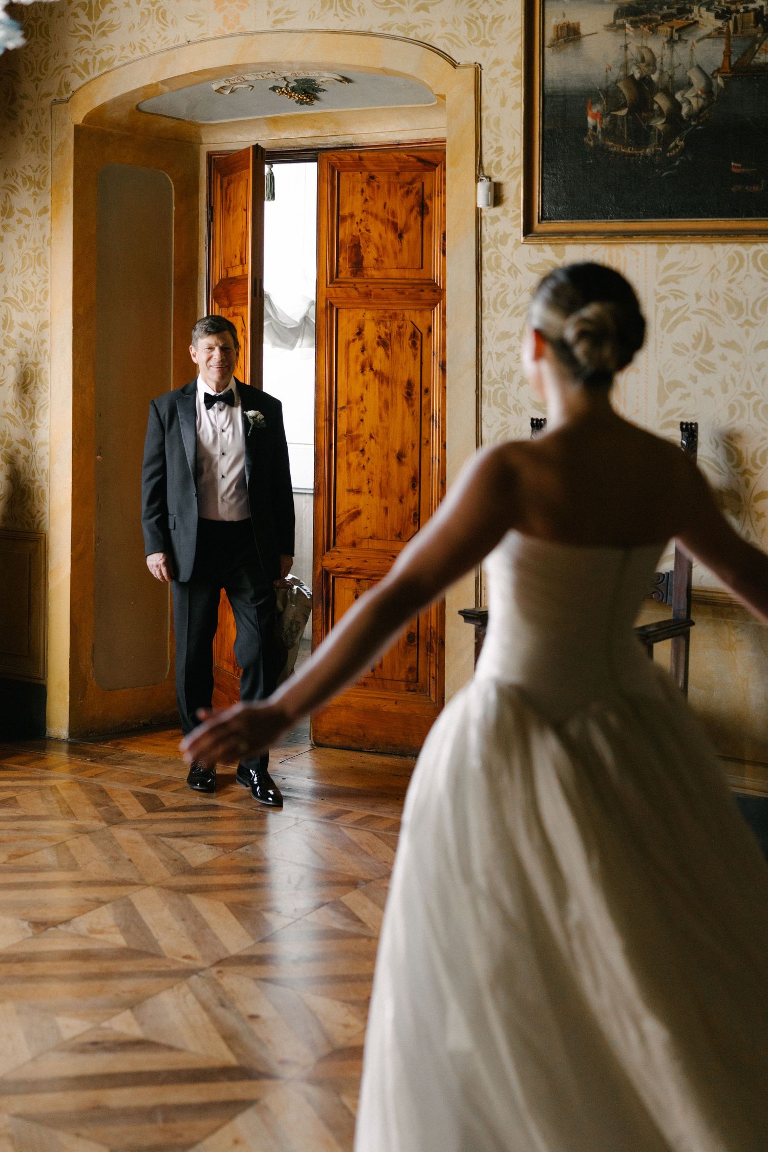 Bride in strapless white gown reaches toward groom in black tuxedo entering ornate wooden doors of elegant venue.