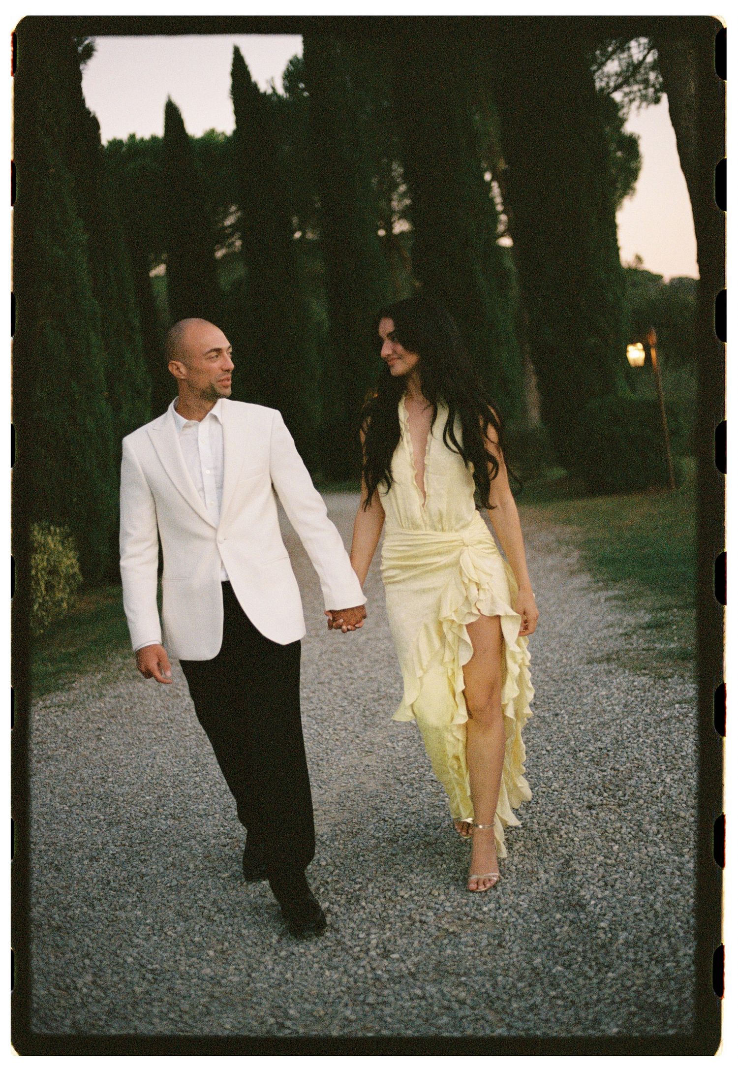 Couple walks hand-in-hand on gravel path lined with cypress trees, man in white blazer, woman in yellow dress.