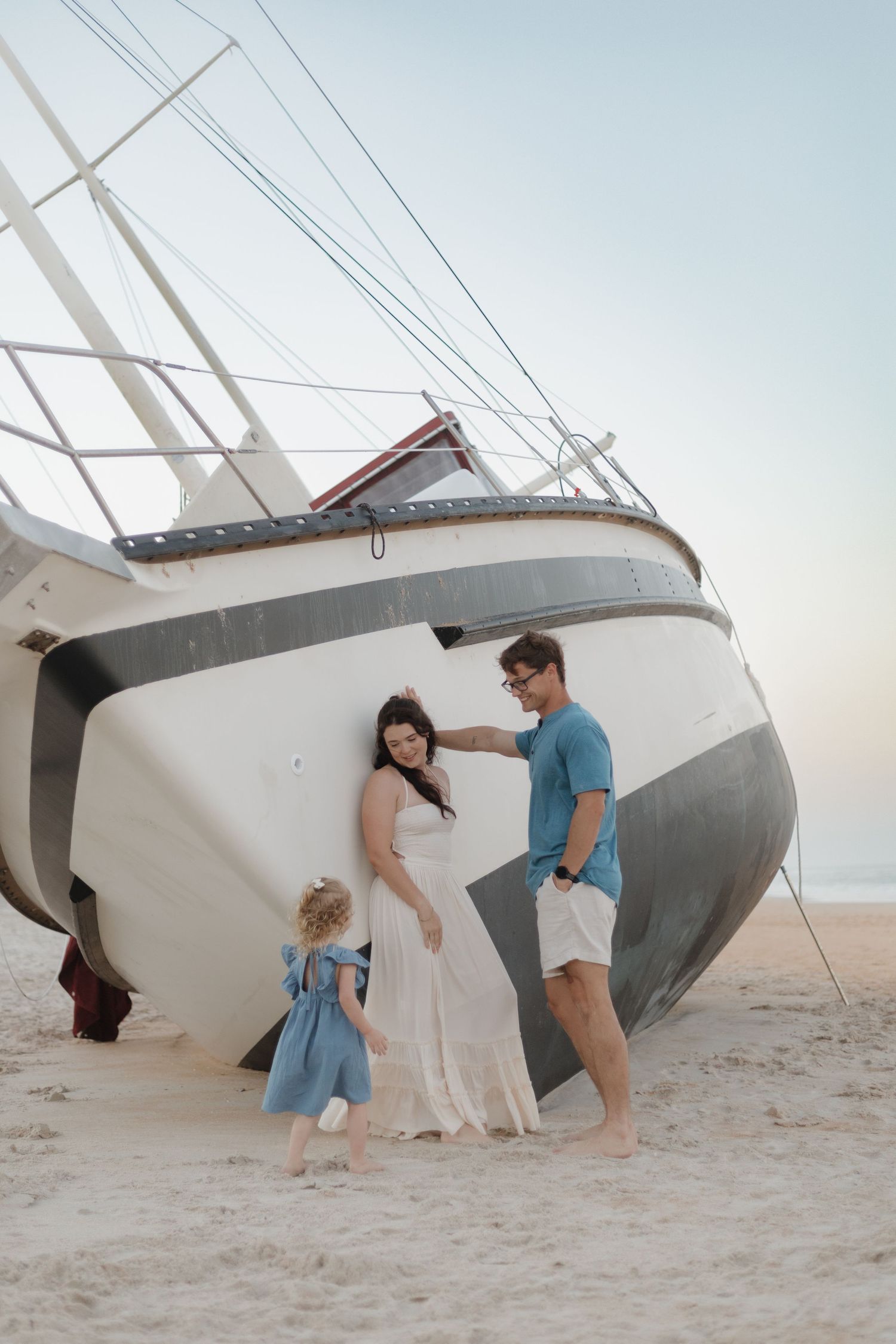 Family posing beside a beached sailboat on sandy shore, woman in white dress, man in blue shirt, young girl in blue dress.