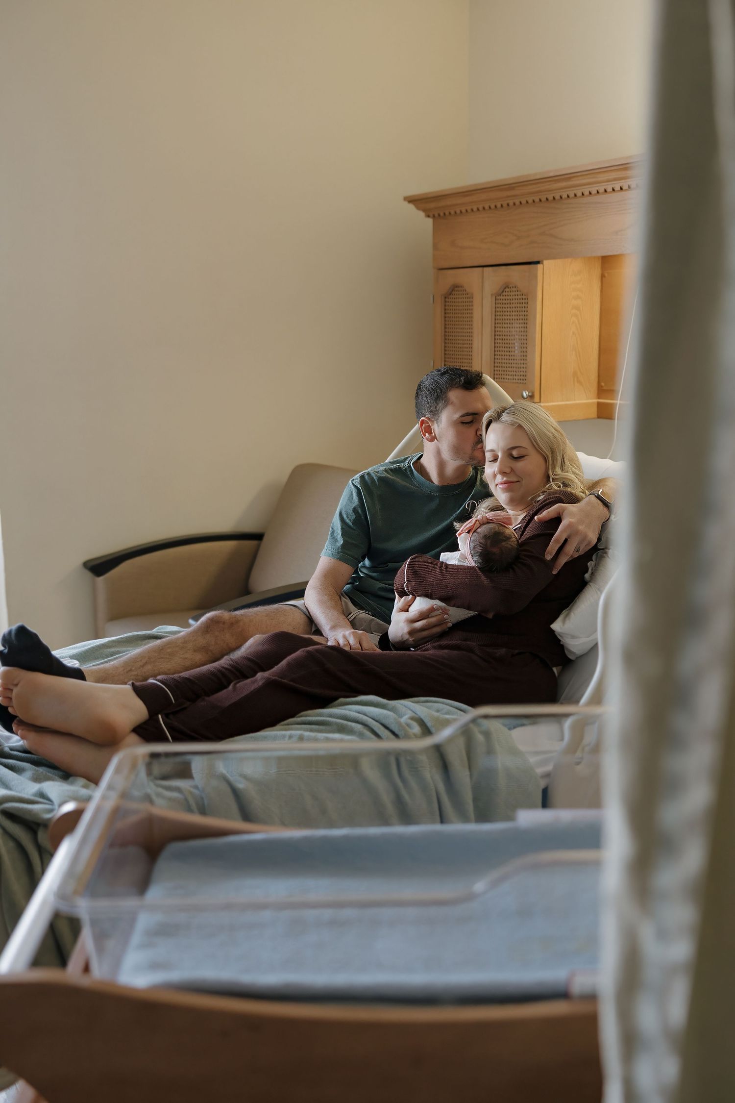 New parents sit together on hospital bed, tenderly cradling their newborn baby in a warm maternity room.