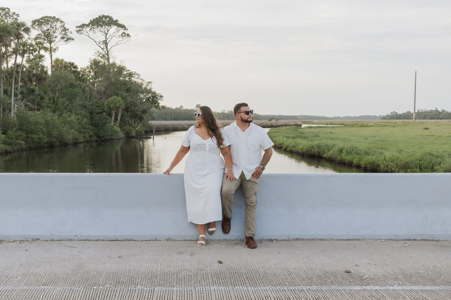 Couple in white outfits standing on a bridge overlooking a serene marsh waterway surrounded by lush greenery.