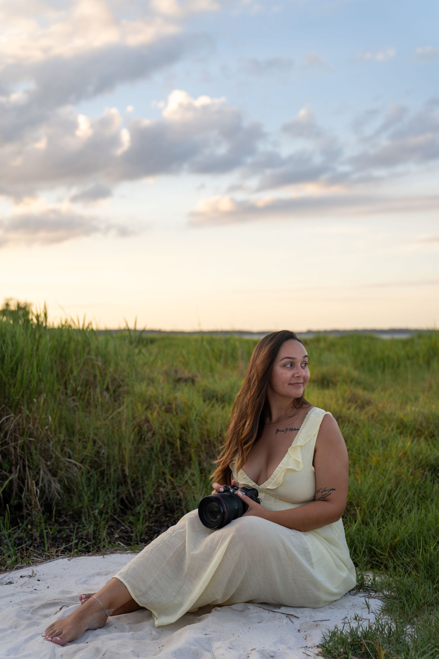 Woman in yellow dress sitting on sand holding camera at sunset with green marsh grass and cloudy sky.
