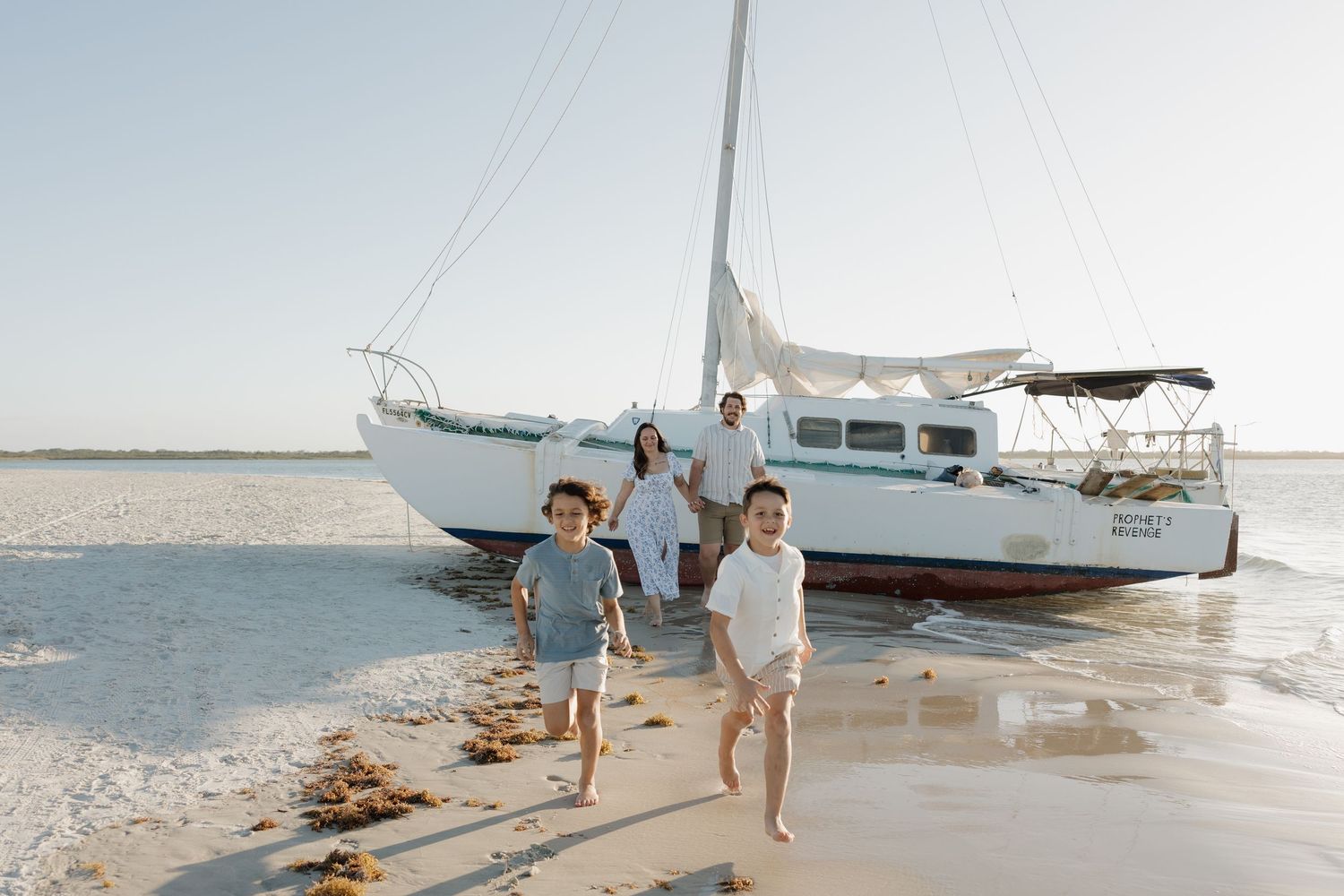 Family of four walking on sandy beach from white catamaran sailboat on calm clear day.