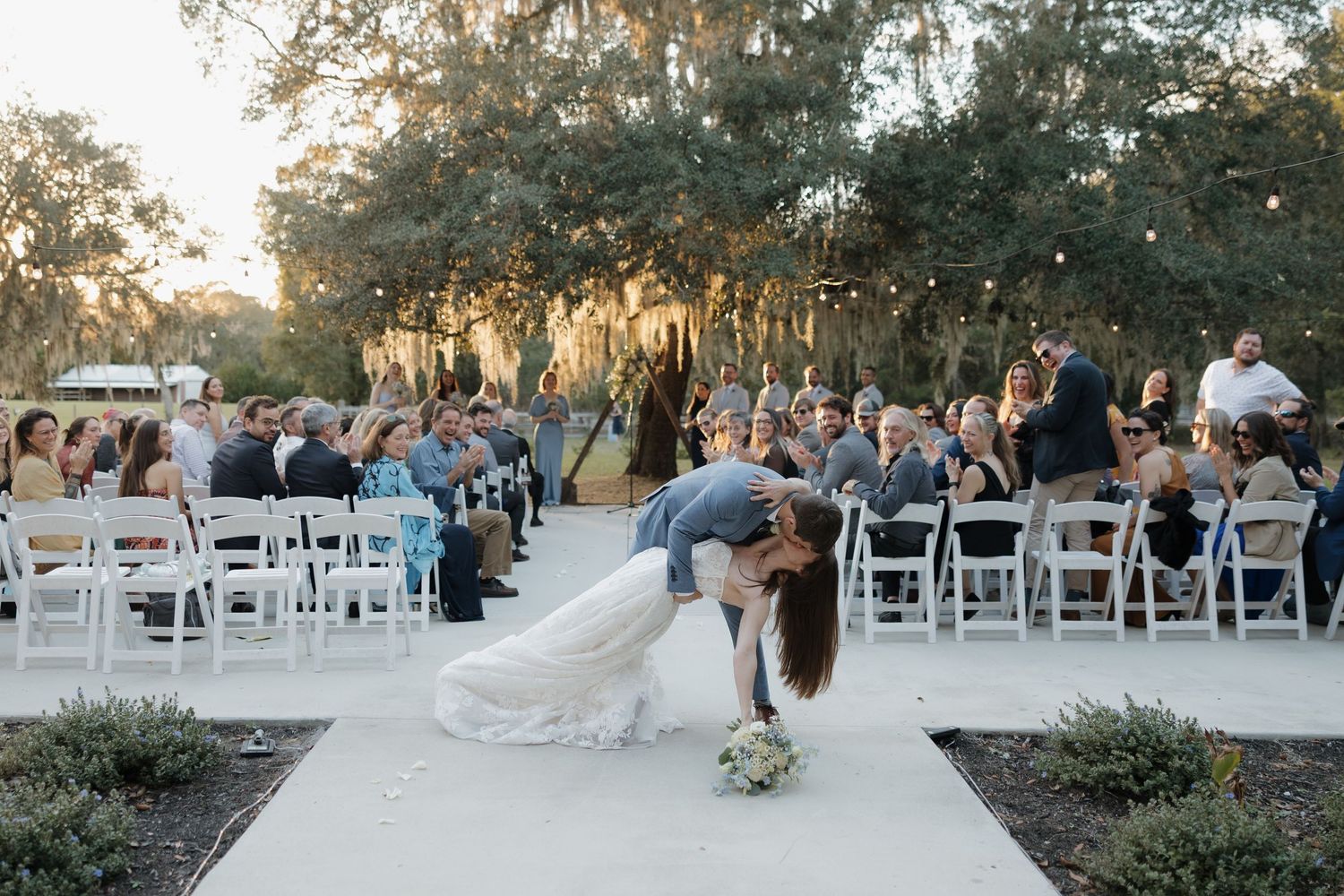 Groom dips bride at outdoor wedding ceremony with white chairs and guests under string lights among trees.