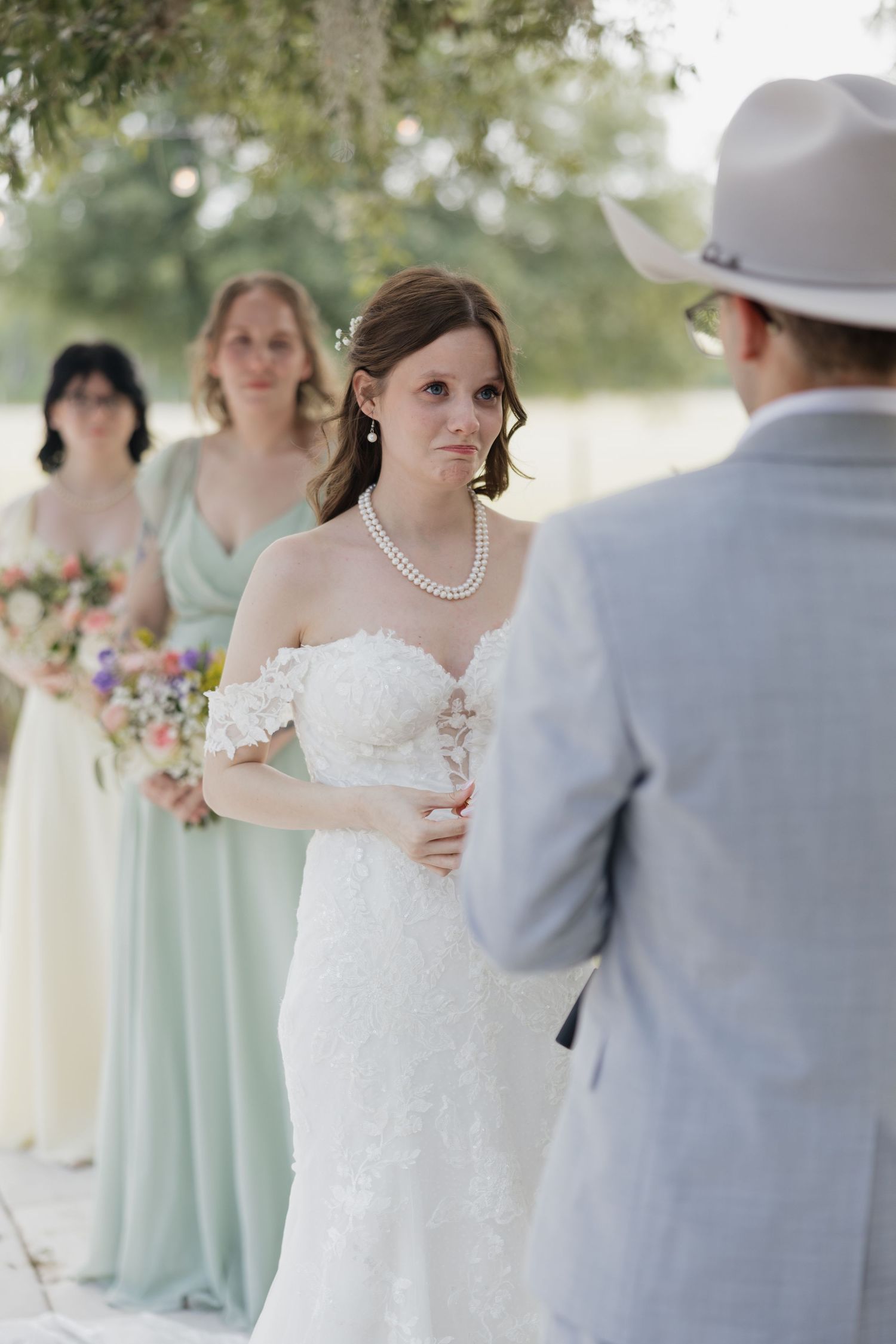 Emotional bride in white lace gown and pearl necklace stands with bridesmaids in mint dresses during outdoor wedding ceremony.