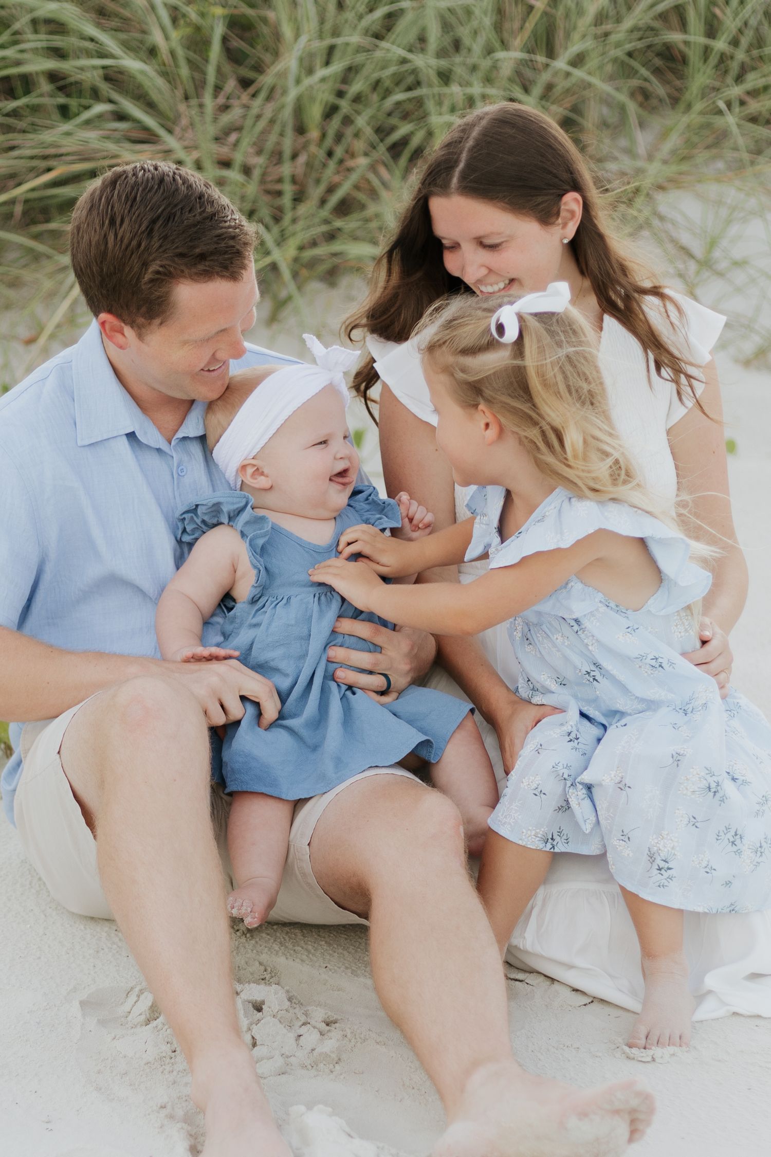 Happy family of four sitting on sandy beach, parents smiling at laughing baby and toddler girl in matching blue dresses.