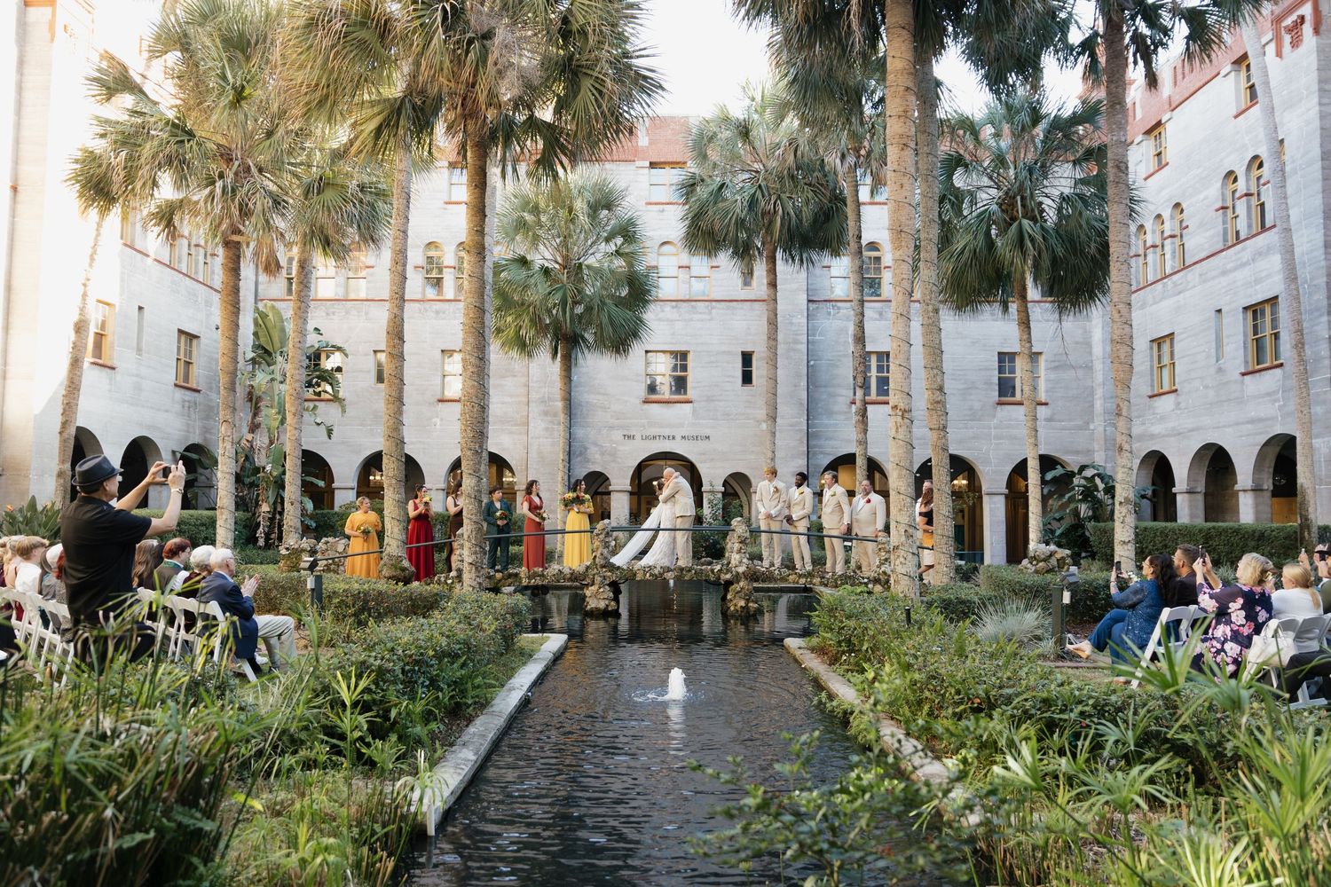 Outdoor wedding ceremony in a lush courtyard with palm trees, reflecting pool, and elegant stone arches at a historic venue.