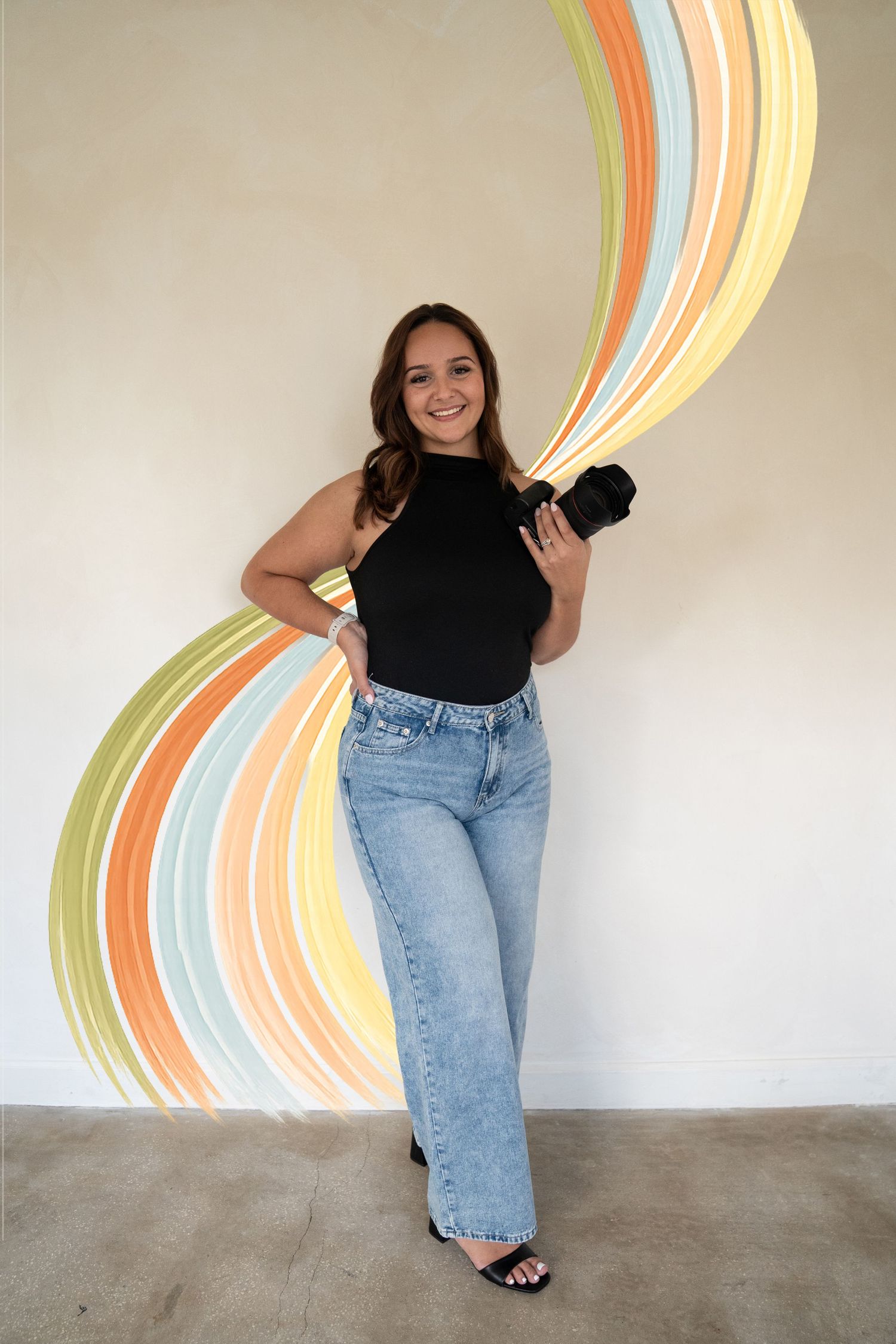 Young woman in black top and jeans holds camera, posing in front of colorful swirl mural on white wall.
