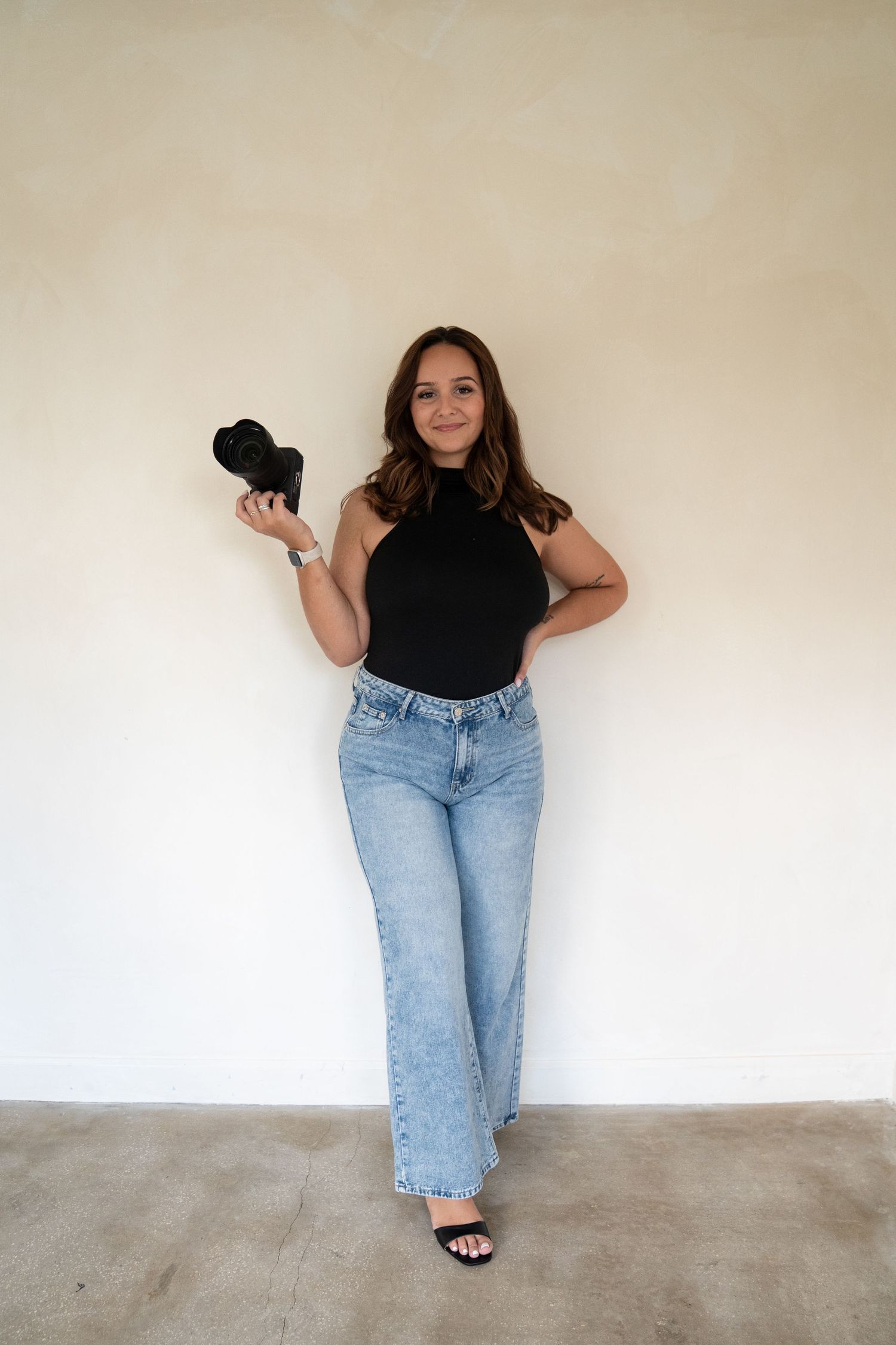 Artistic poses against white wall wearing black top and light denim jeans.