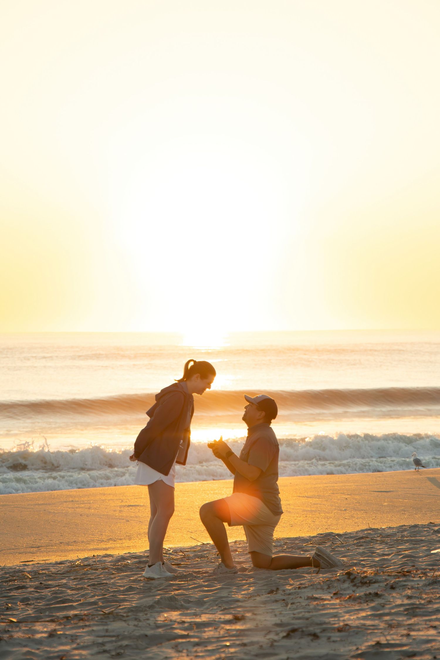 Man proposing on one knee to woman on a sandy beach at golden sunset with ocean waves in the background.