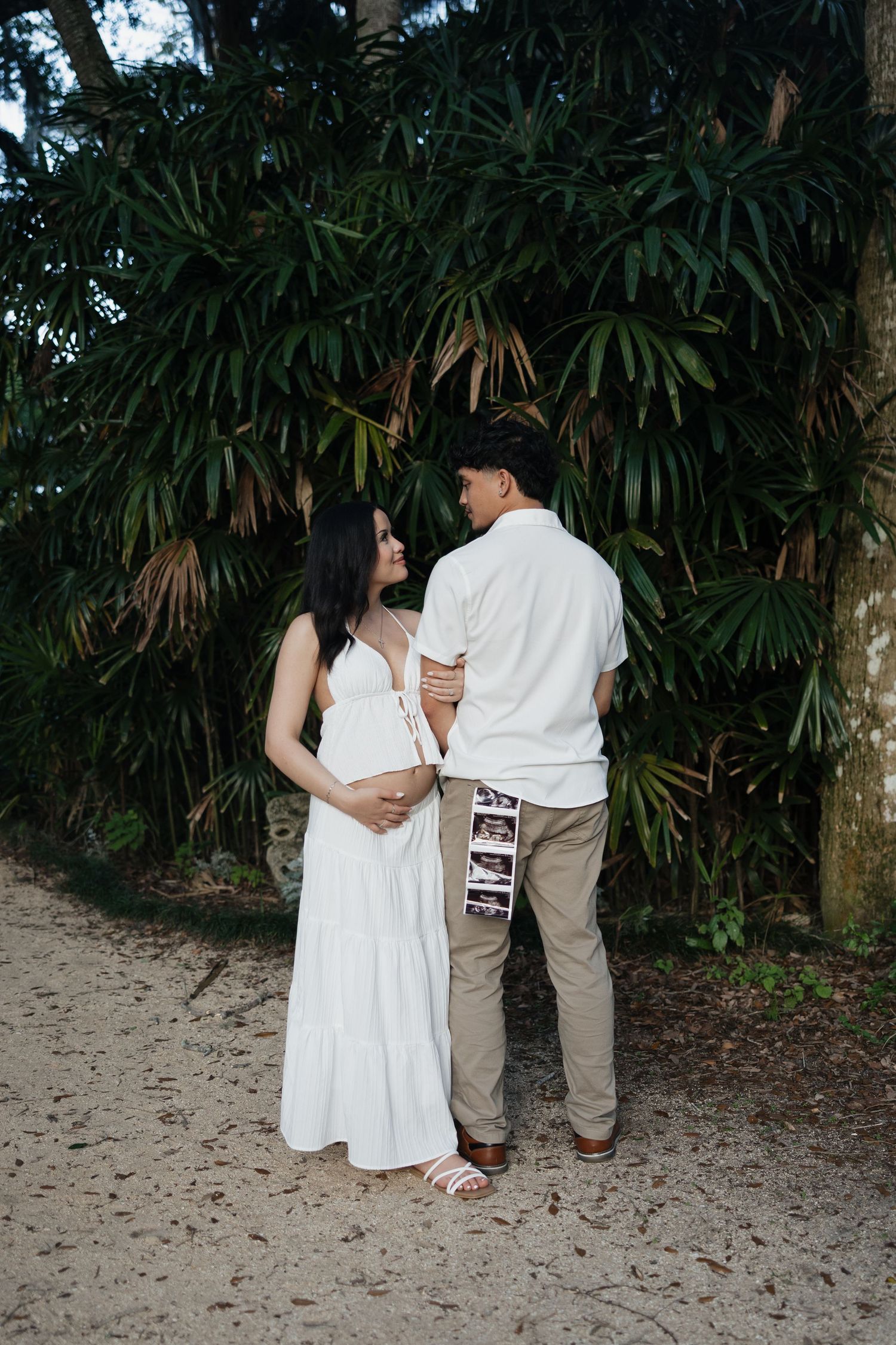 Pregnant couple in white dress and khaki pants holding ultrasound photos, standing on sandy path with tropical trees.