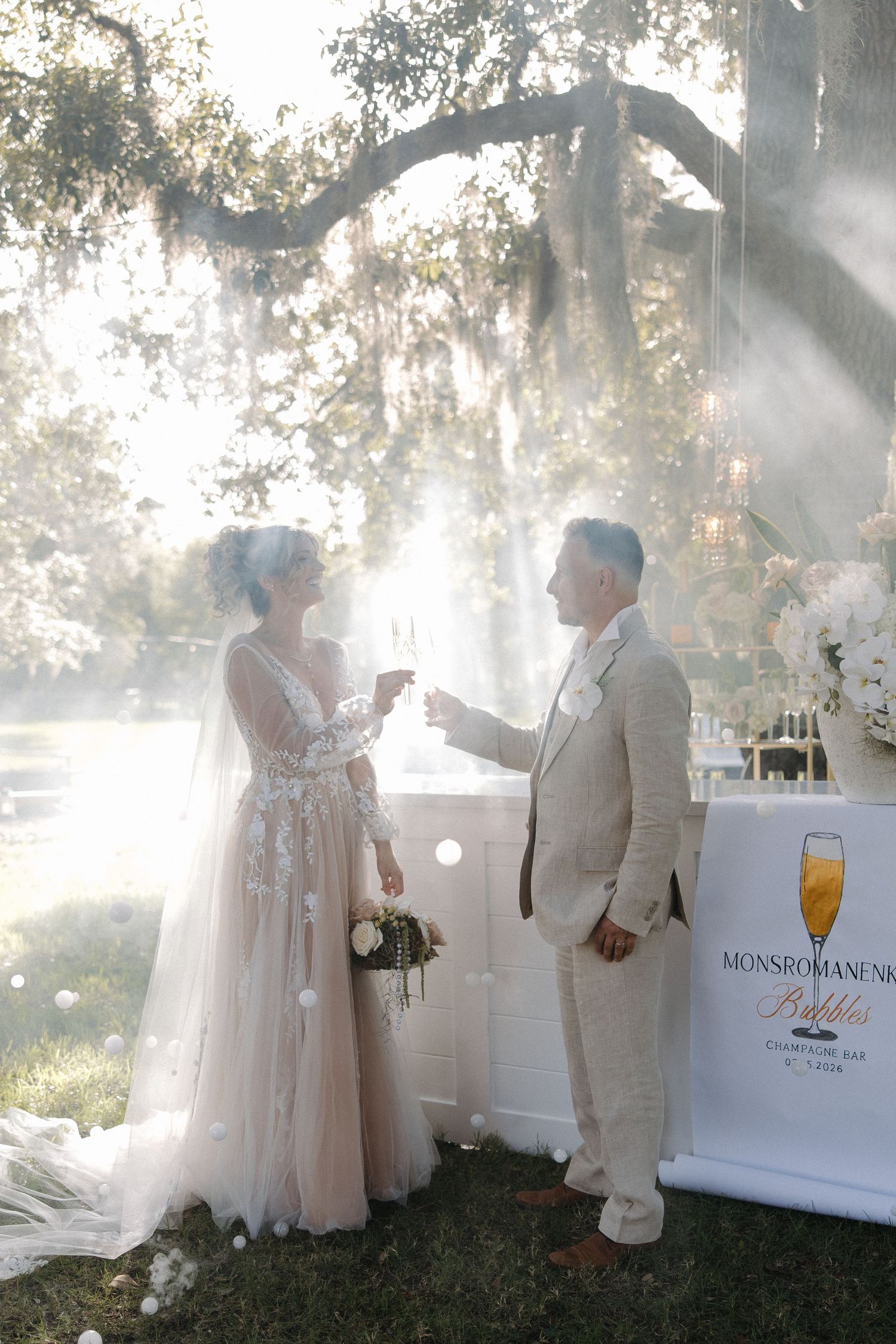 Bride and groom toasting champagne at outdoor wedding by Mons Romane bubbles bar under misty oak trees.