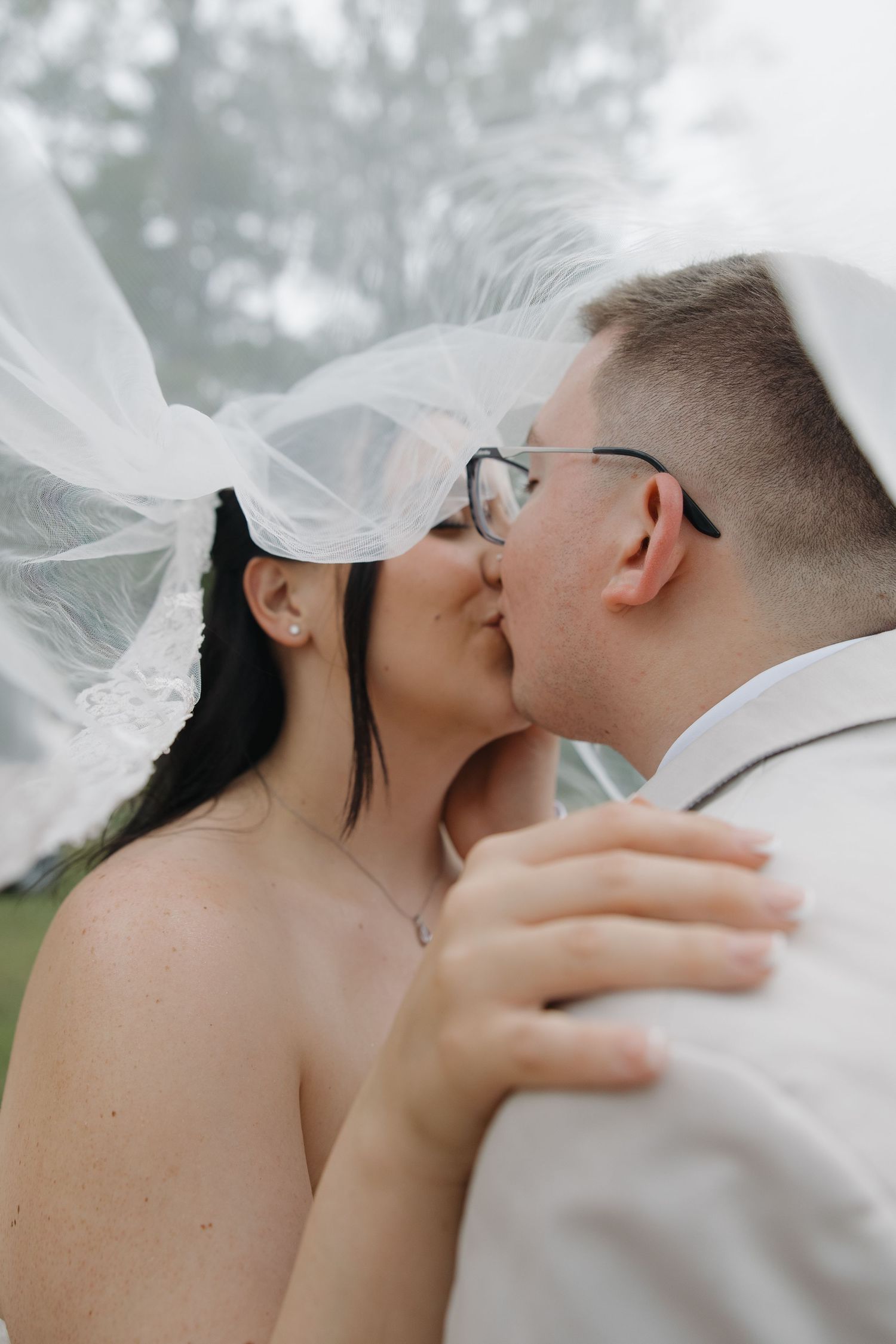 A bride and groom share a romantic kiss under a flowing white veil on their wedding day outdoors.