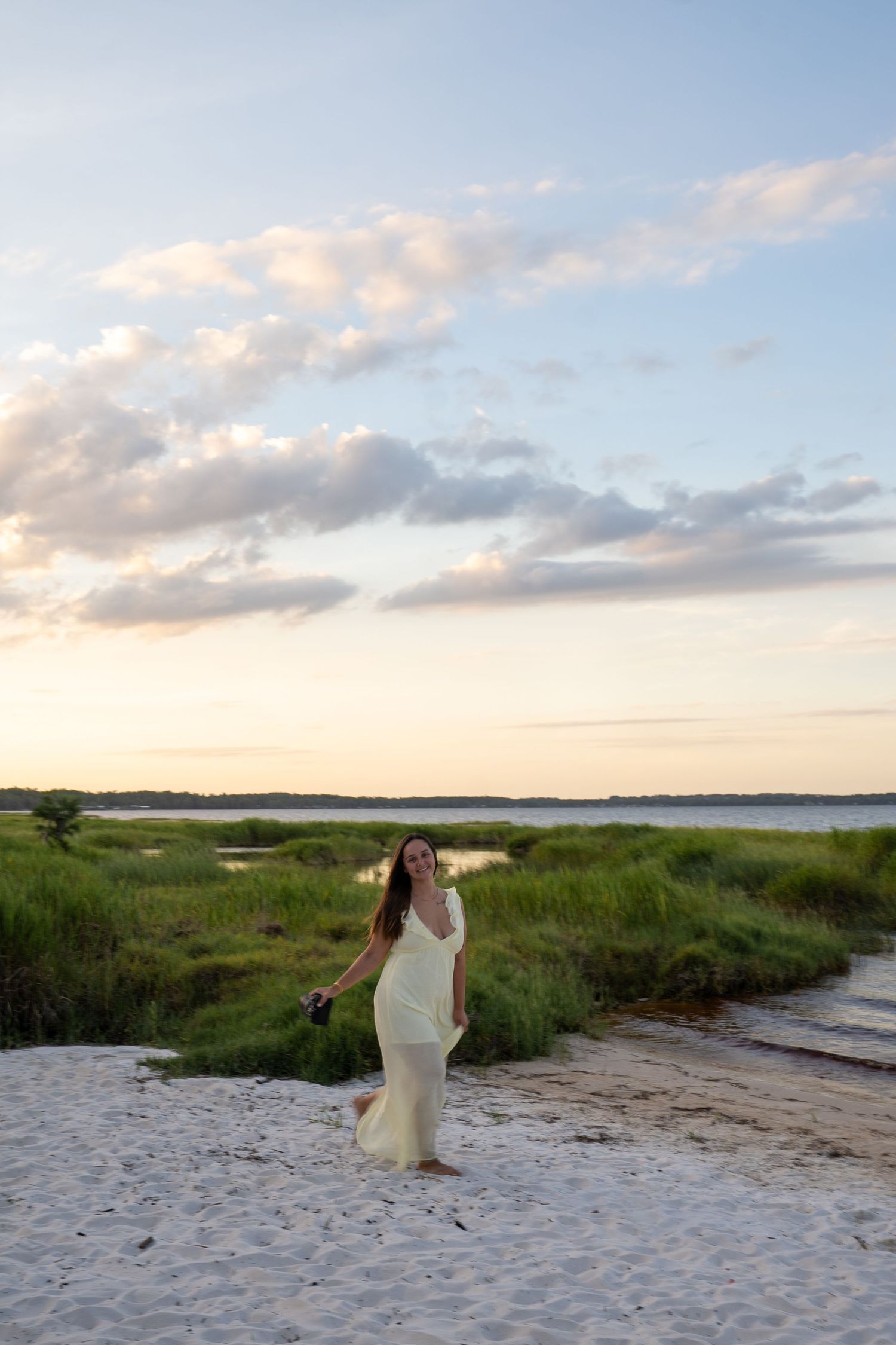 Woman in white dress walking on sandy beach path at sunset with marsh grass and calm water in background.