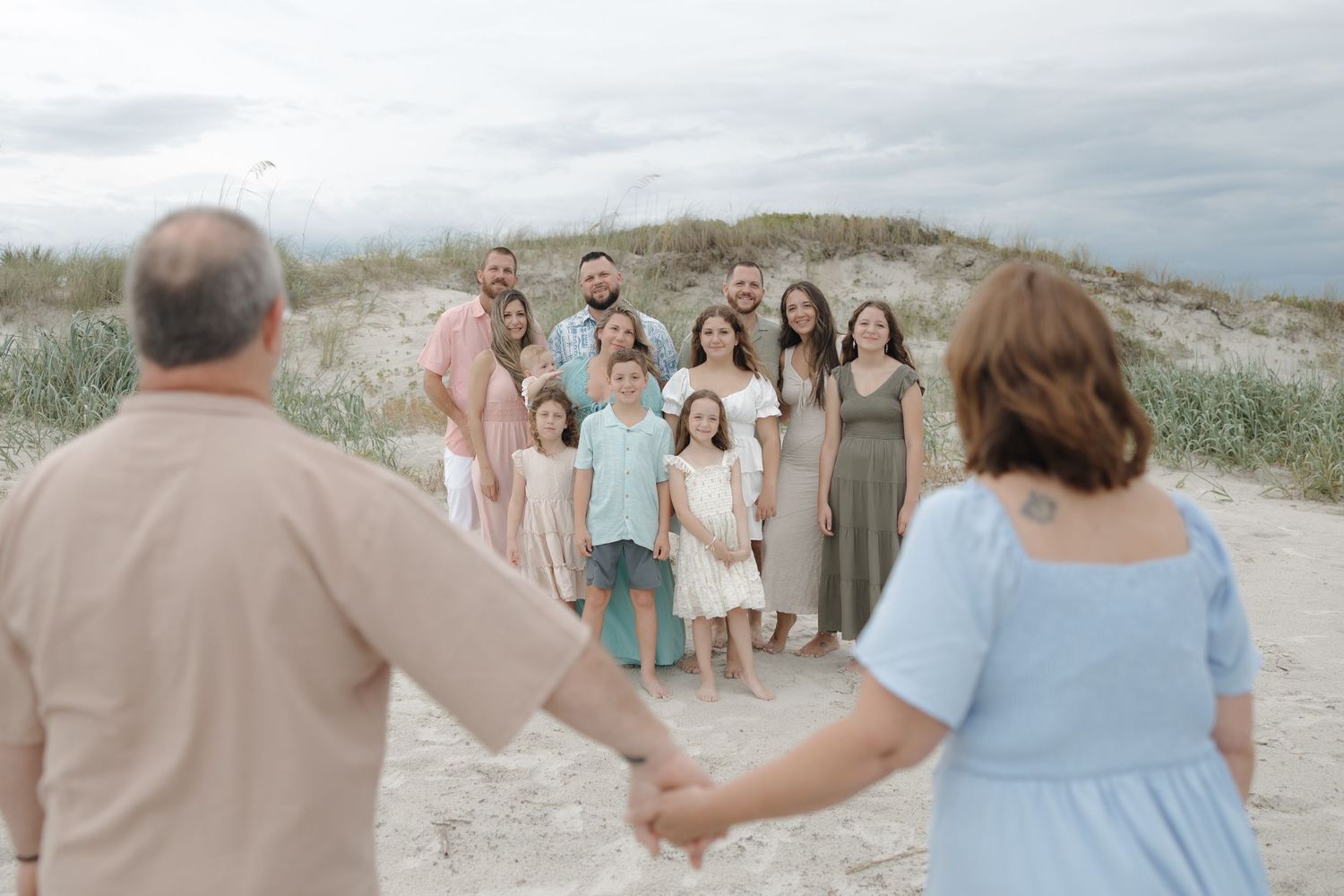 Large family group photo on sandy beach dunes, with couple holding hands in foreground facing the group.