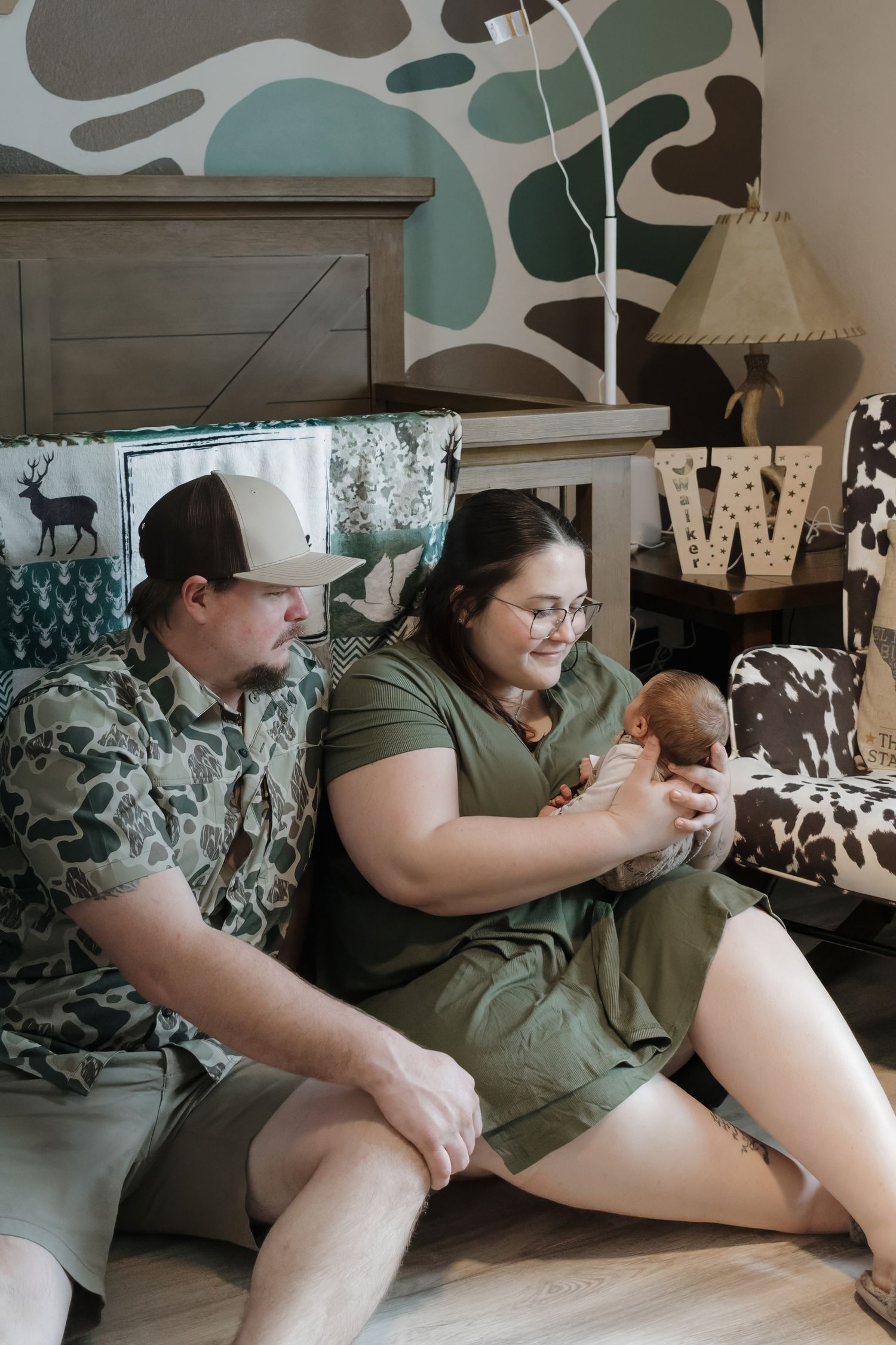 A couple sits on the floor holding their newborn baby in a camo-themed nursery with a wooden W letter decor.