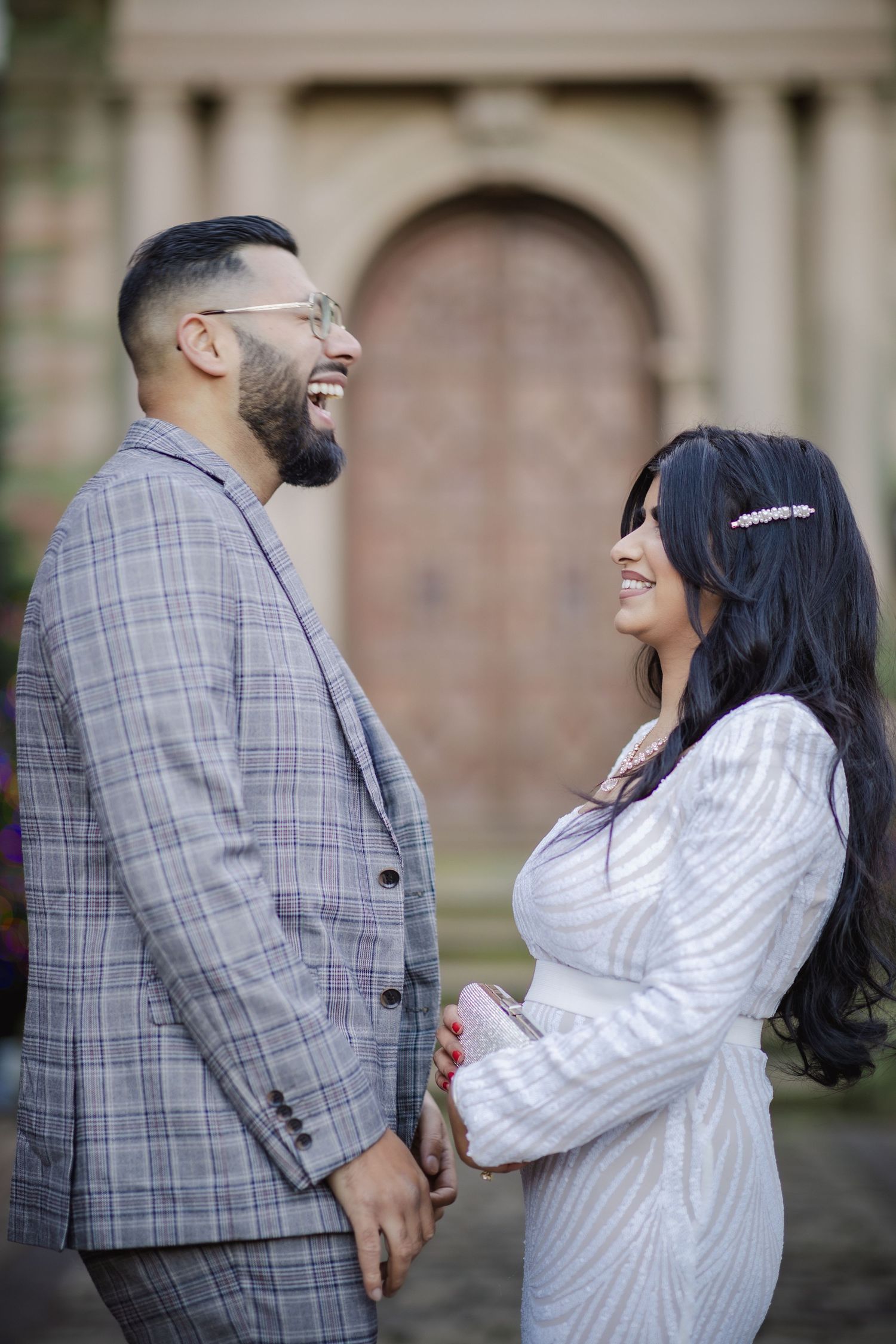 A laughing couple in formal attire stands together outdoors, the man in a plaid suit and woman in white dress.