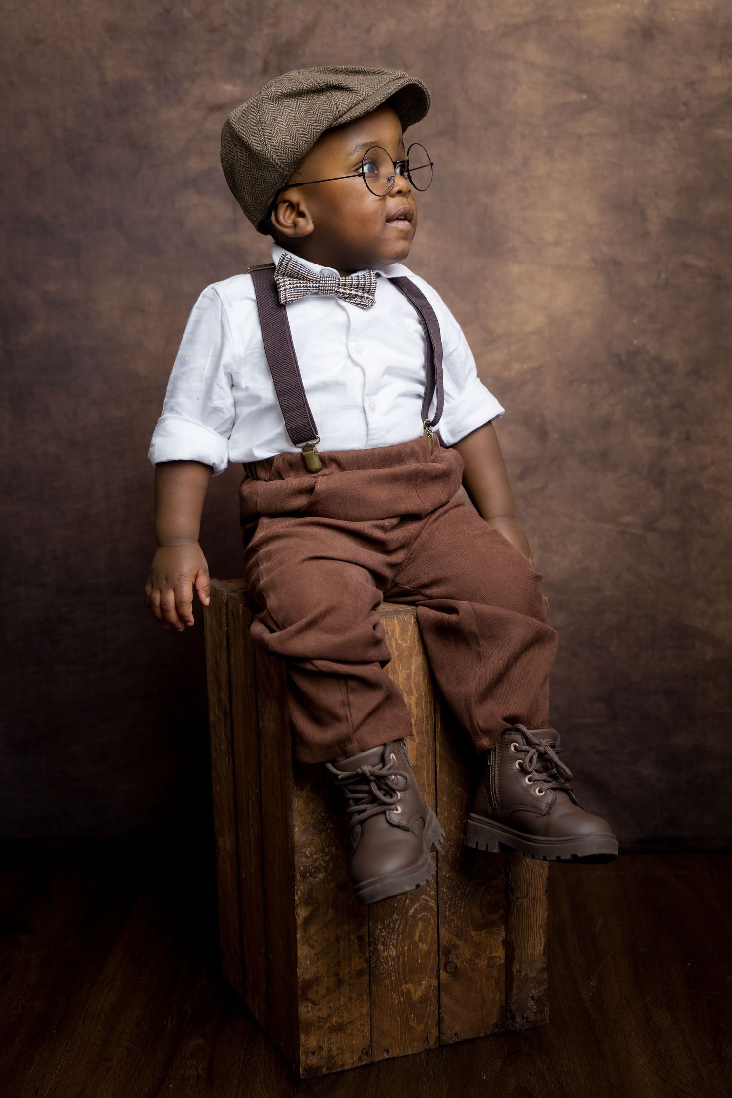 Adorable toddler boy in vintage outfit with suspenders, bow tie, flat cap, glasses, and brown boots sitting on wooden crate.