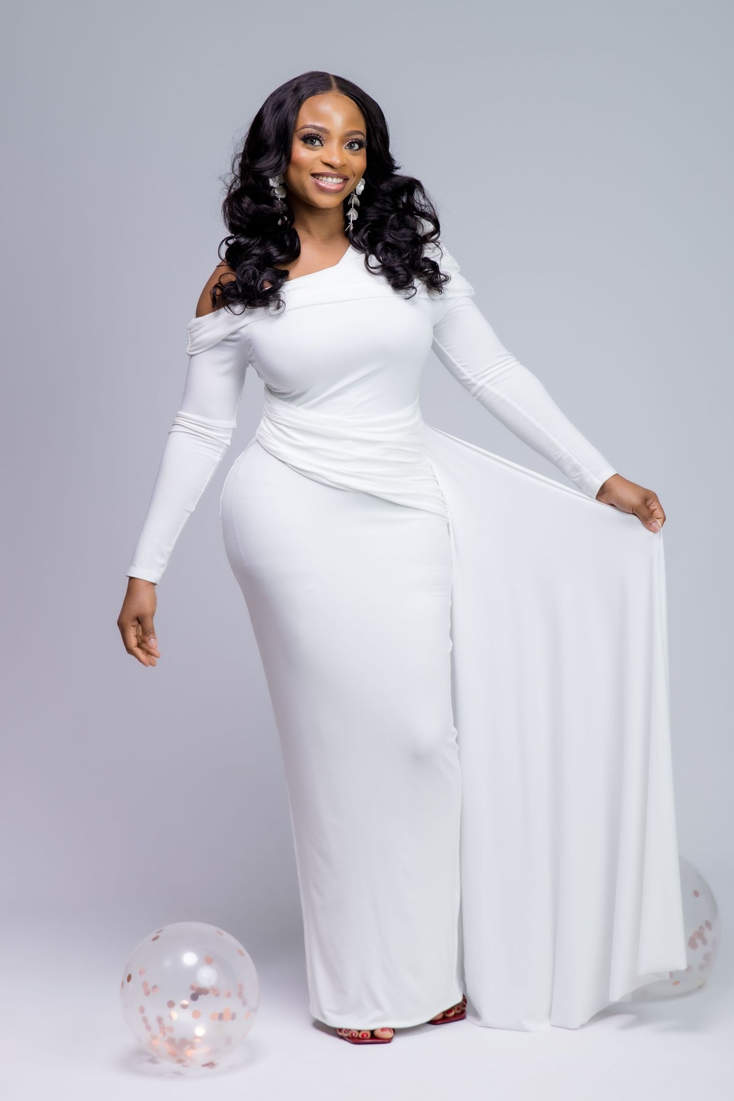 A smiling woman in an elegant white cold-shoulder maxi dress with long sleeves poses gracefully in a studio setting.
