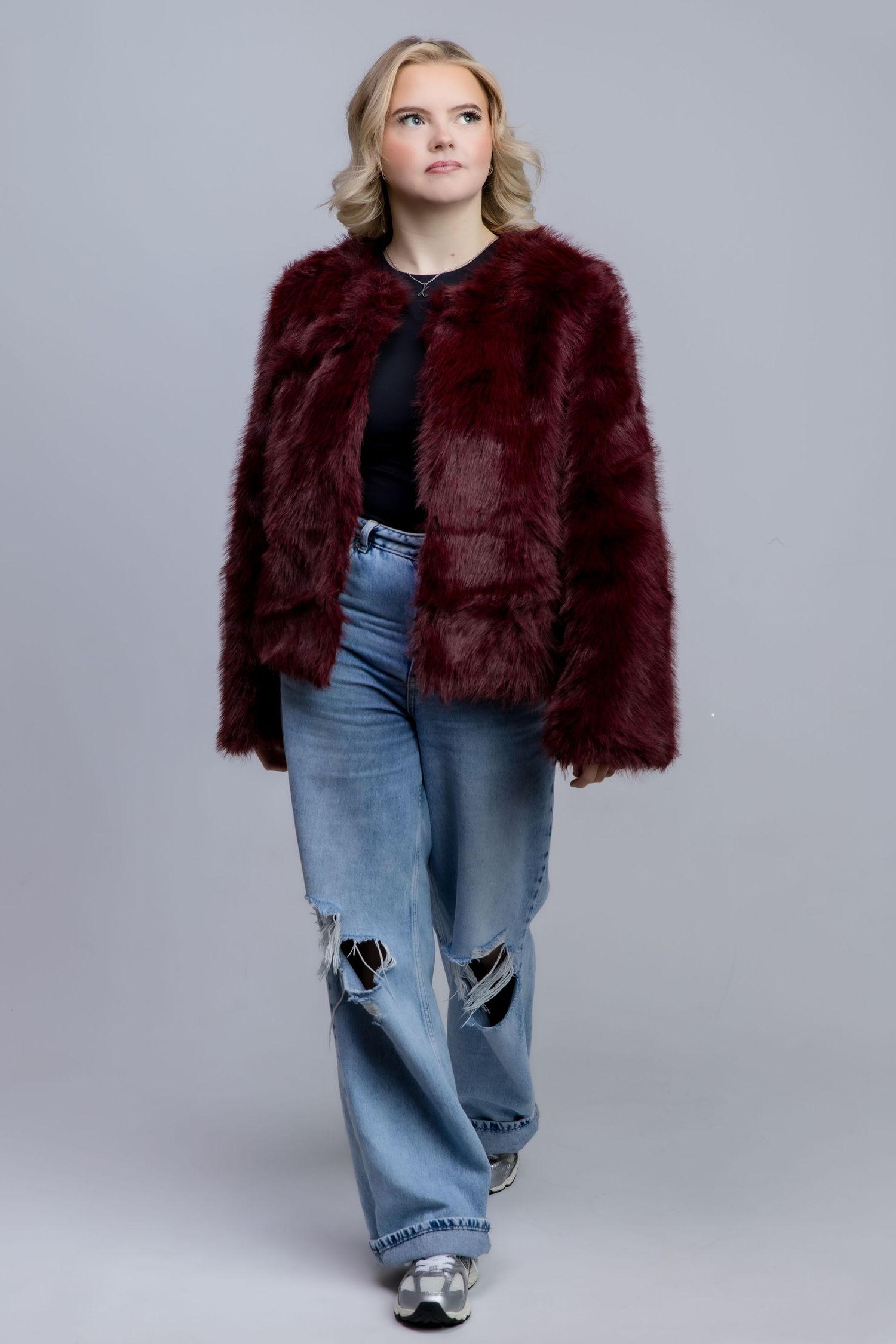 Blonde woman wearing a burgundy faux fur coat with wide-leg ripped jeans and sneakers on a gray background.