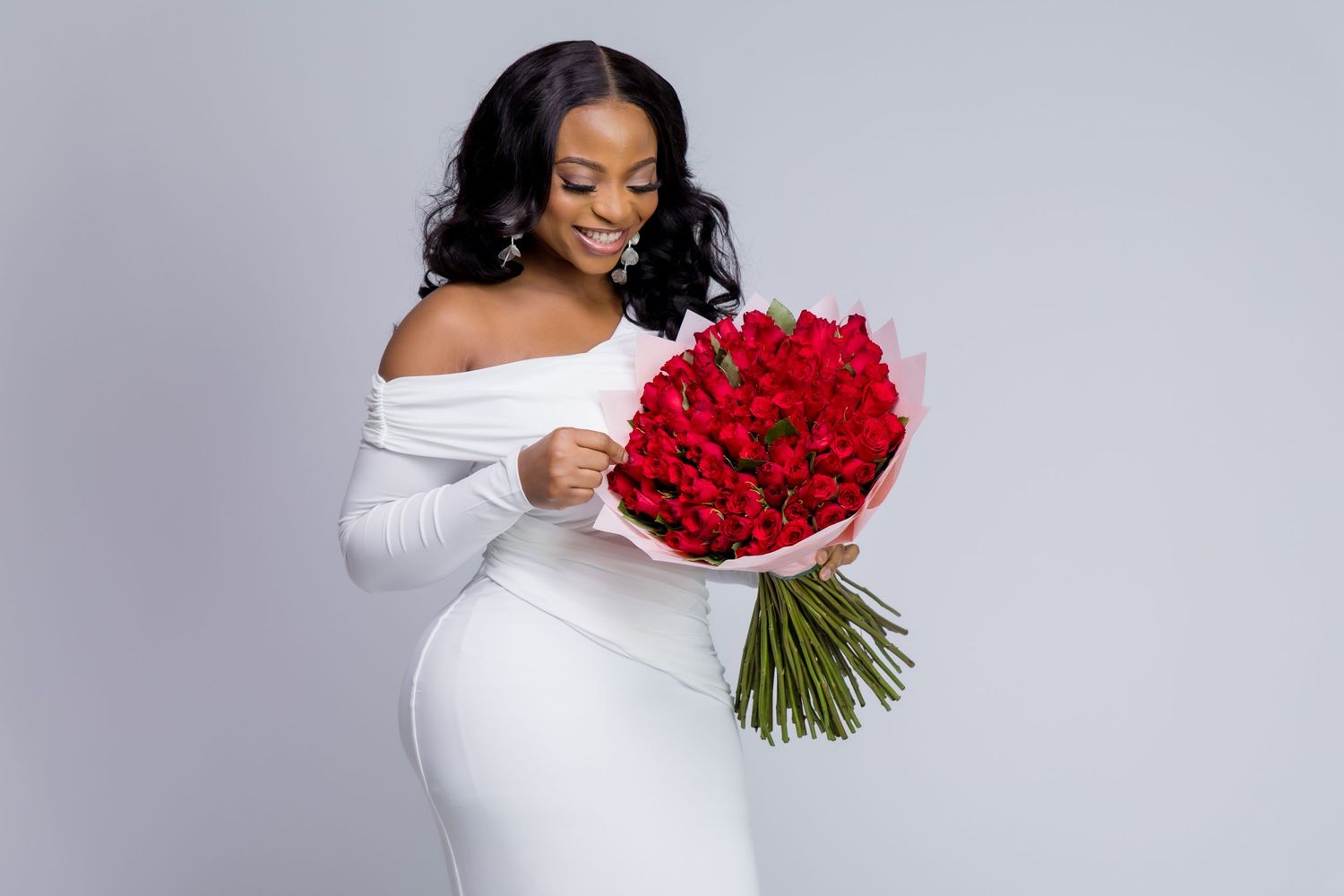 Woman in white off-shoulder dress holding large red rose bouquet, smiling elegantly against gray background.