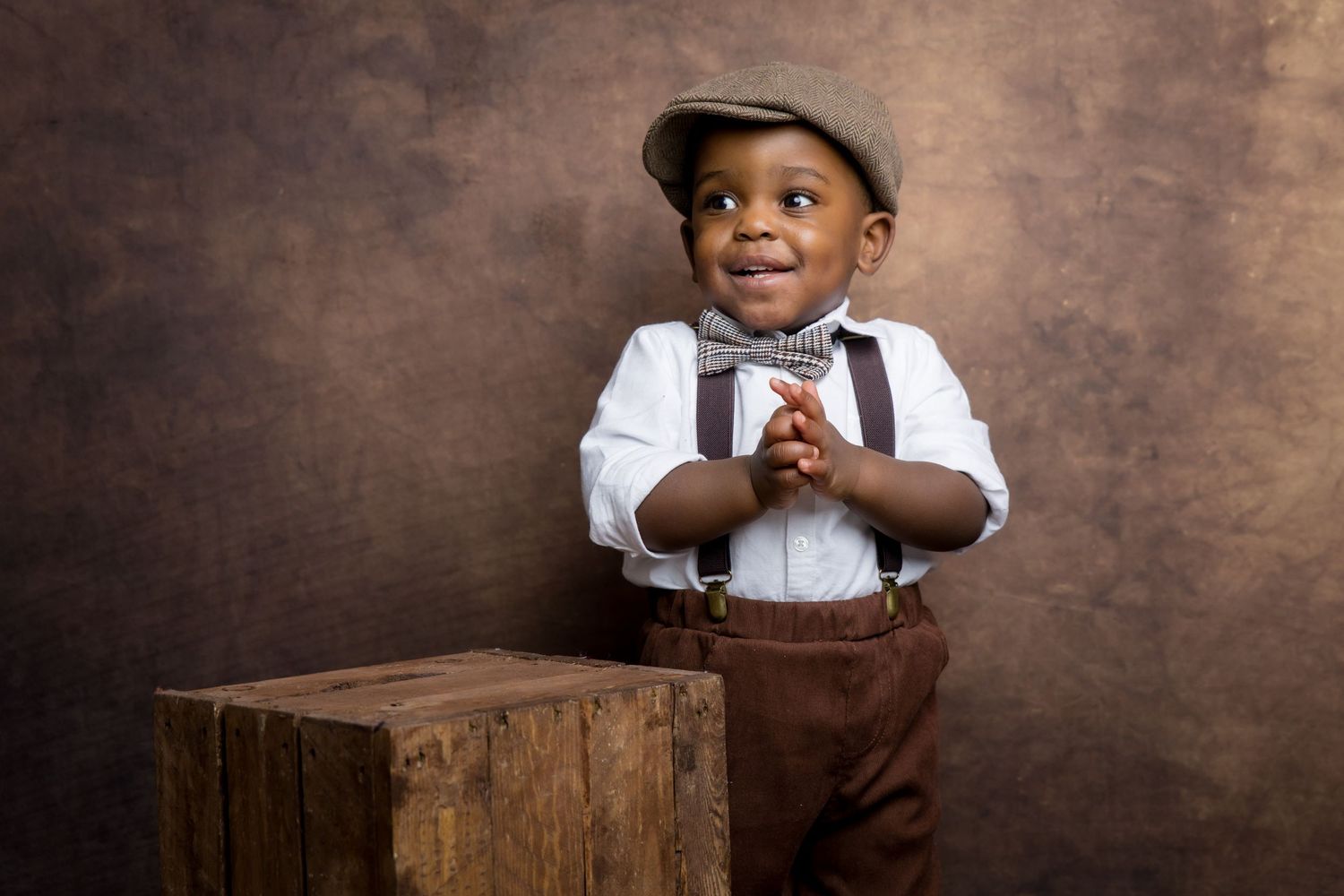 Adorable toddler boy in suspenders, bow tie, and flat cap posing beside a wooden crate on brown backdrop.