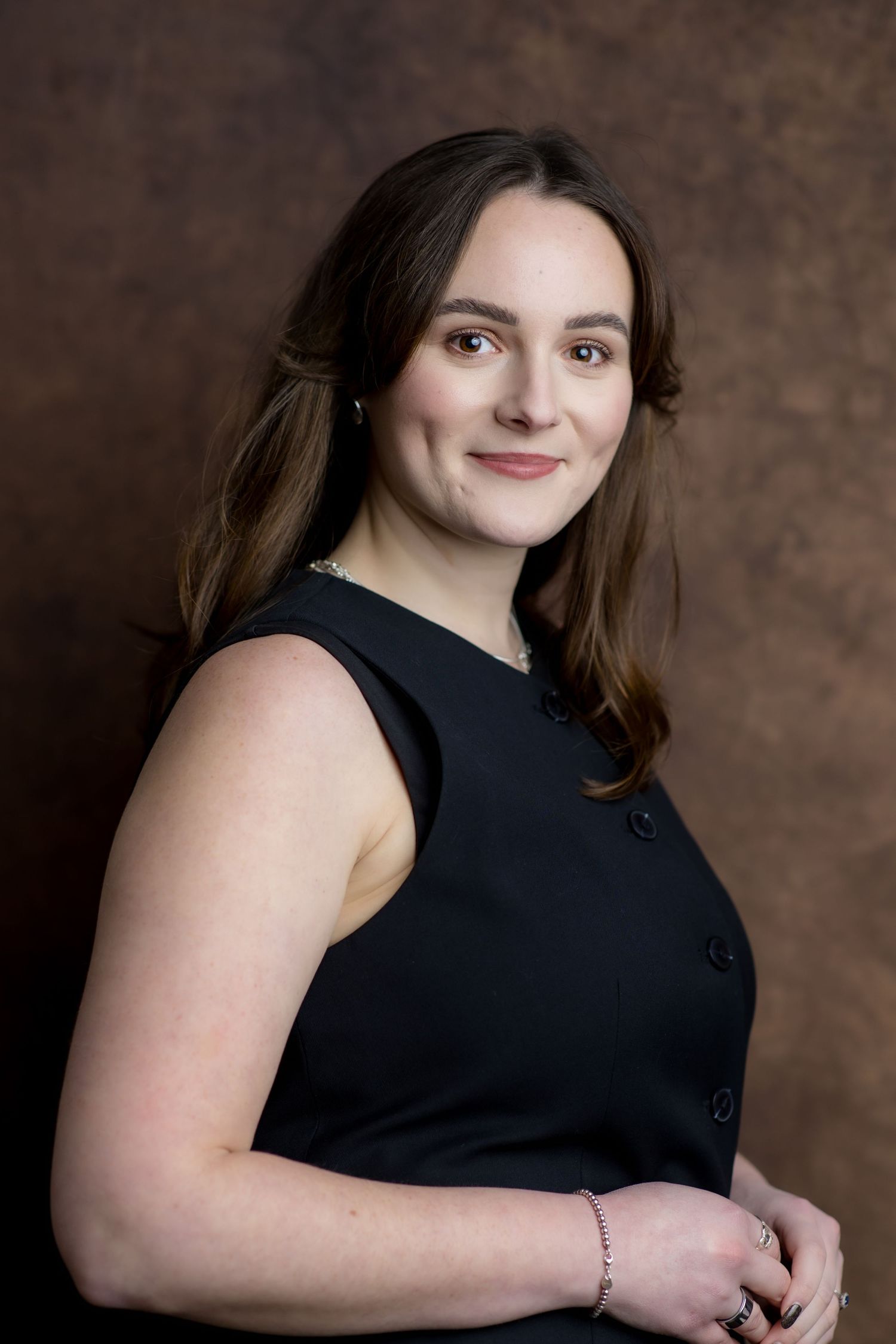 Professional woman in black sleeveless dress smiling confidently against a brown studio background.
