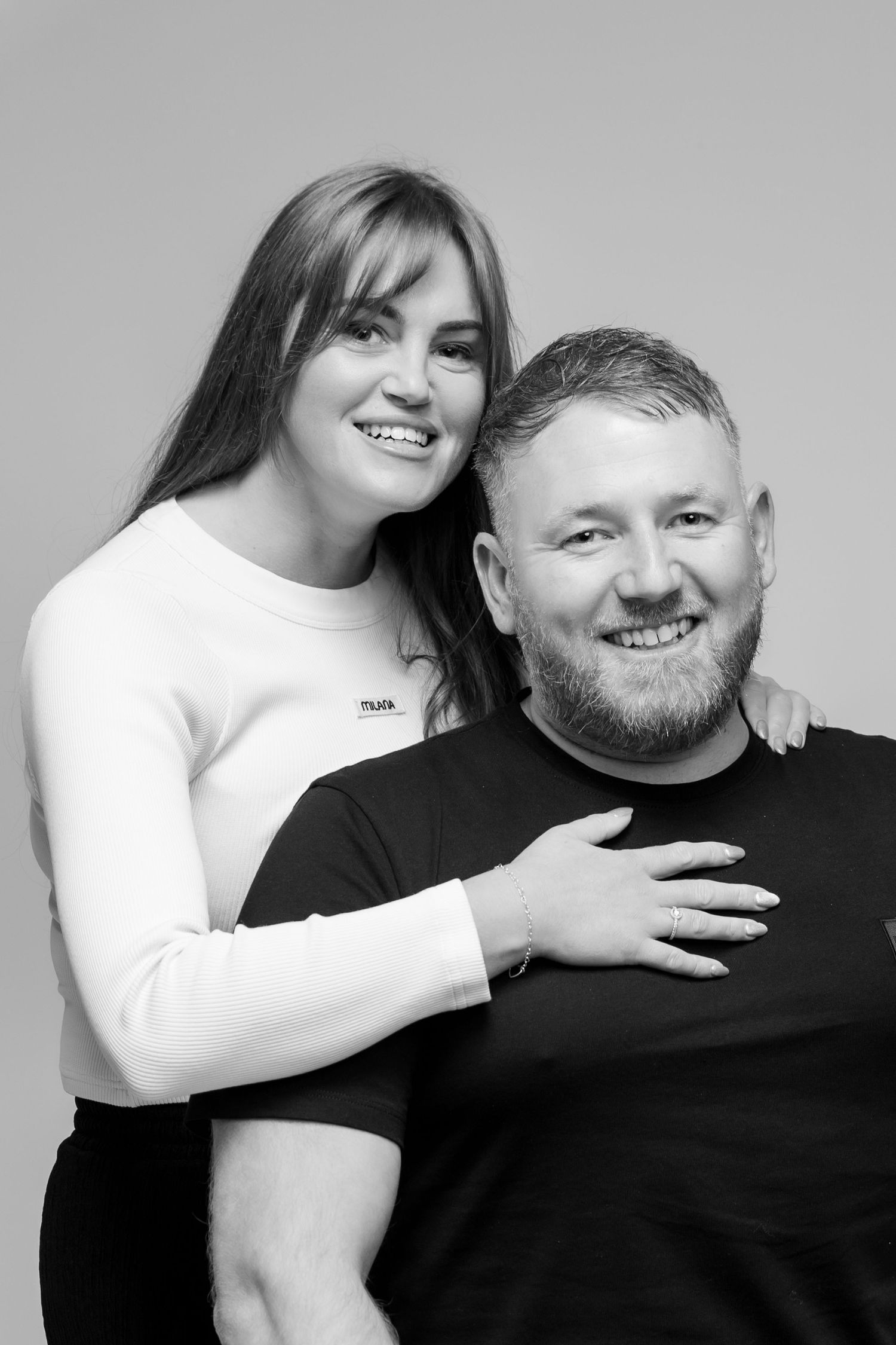 Black and white studio portrait of a smiling couple, woman in white top embracing man in black t-shirt.