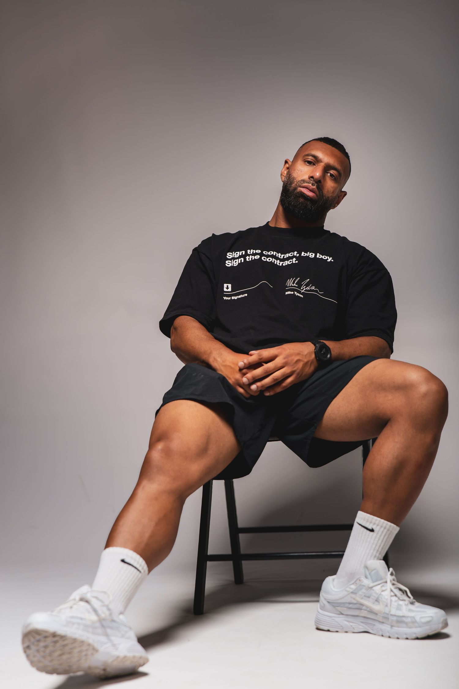 A bearded Black man sits on a stool wearing a black graphic tee, black shorts, white Nike socks, and white sneakers.