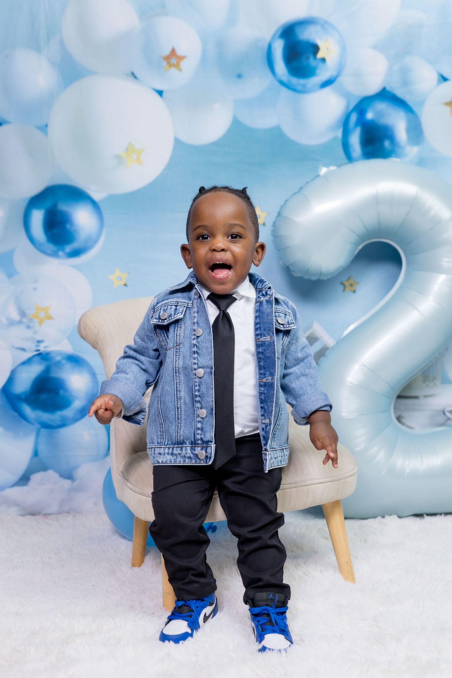 Toddler boy in denim jacket and tie stands before blue balloon backdrop with number 2, celebrating his 2nd birthday.