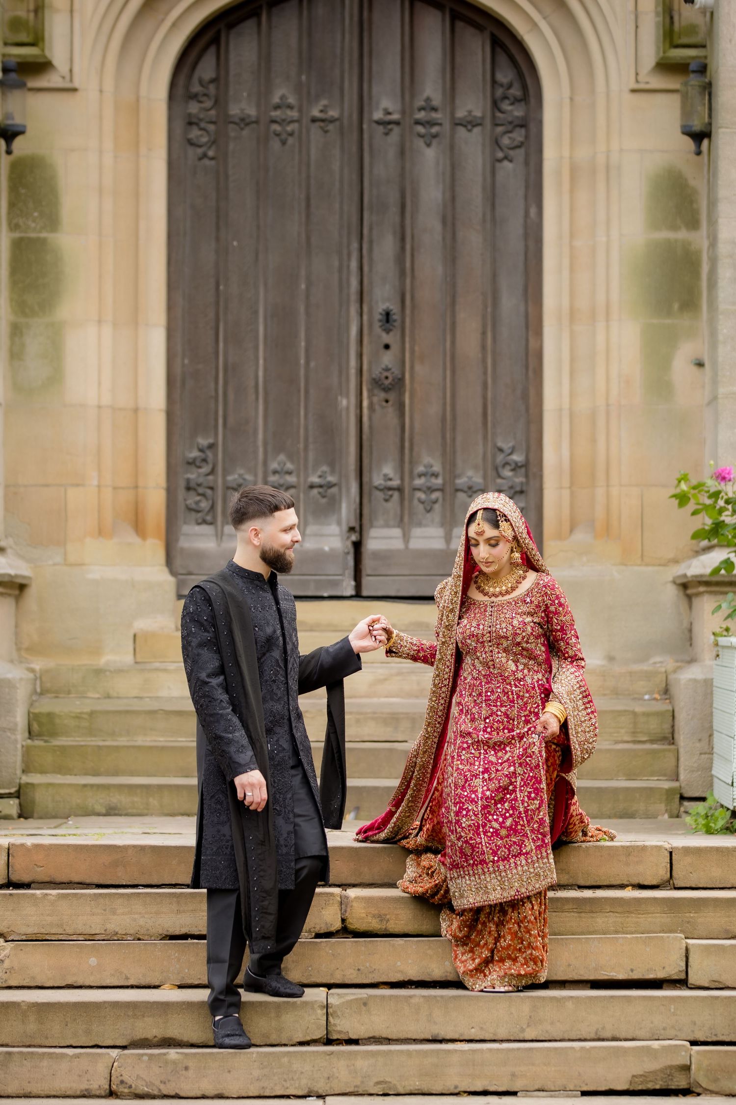 South Asian bride in red lehenga and groom in black sherwani hold hands on stone steps before ornate wooden doors.