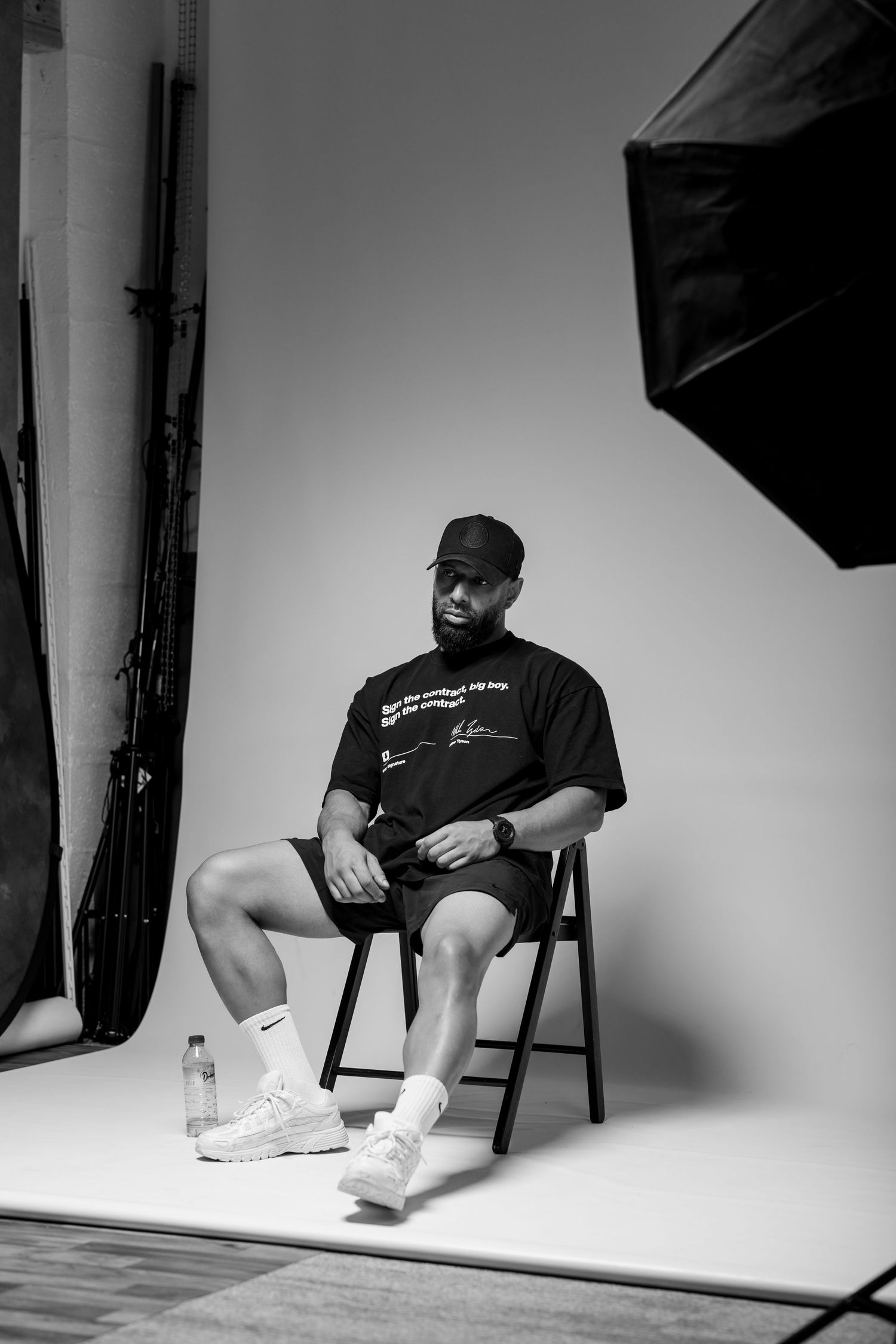 Black and white photo of a man sitting on a folding chair in a photography studio wearing a cap and Nike sneakers.
