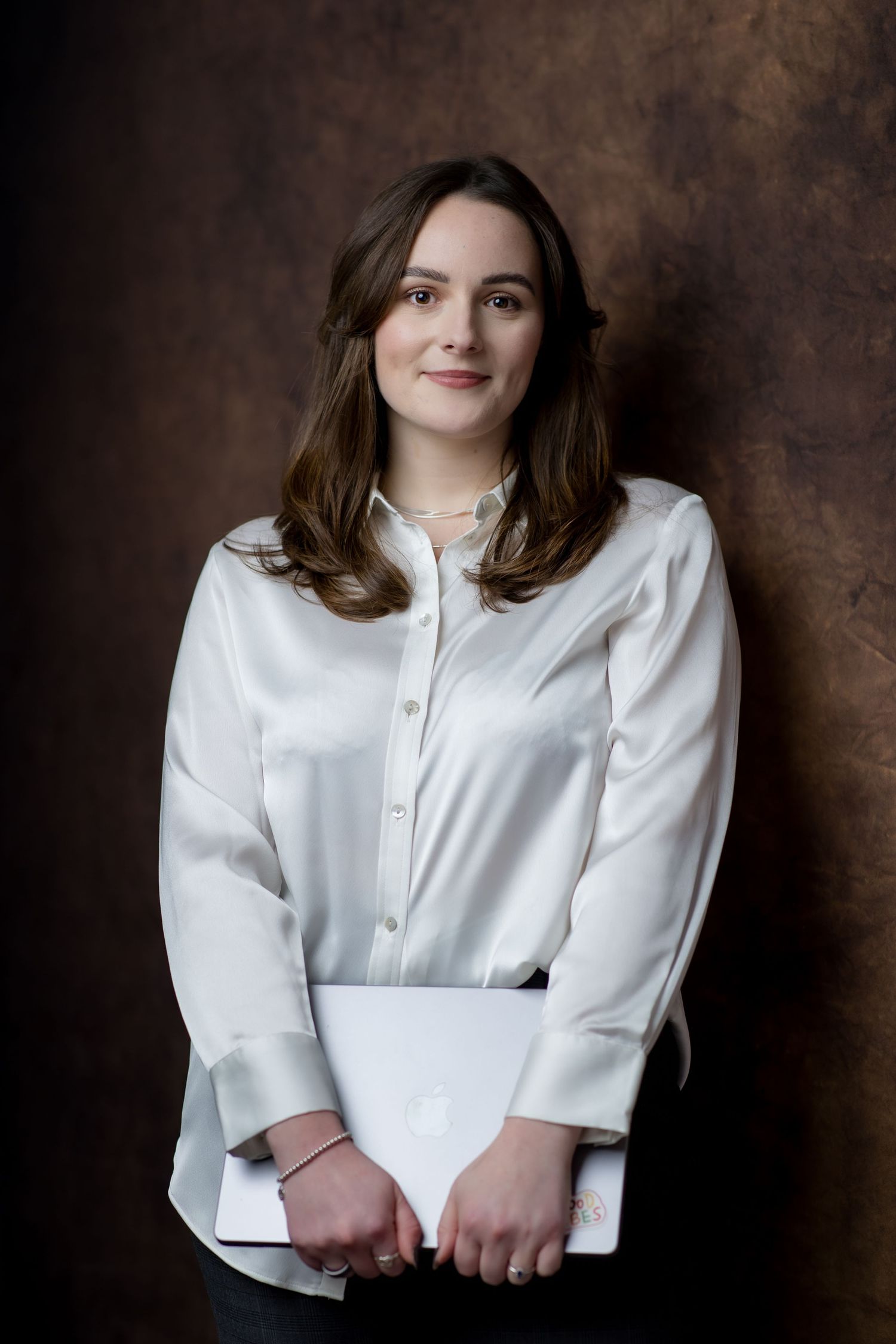 Young professional woman in white satin blouse holding laptop, posing confidently against dark brown studio background.