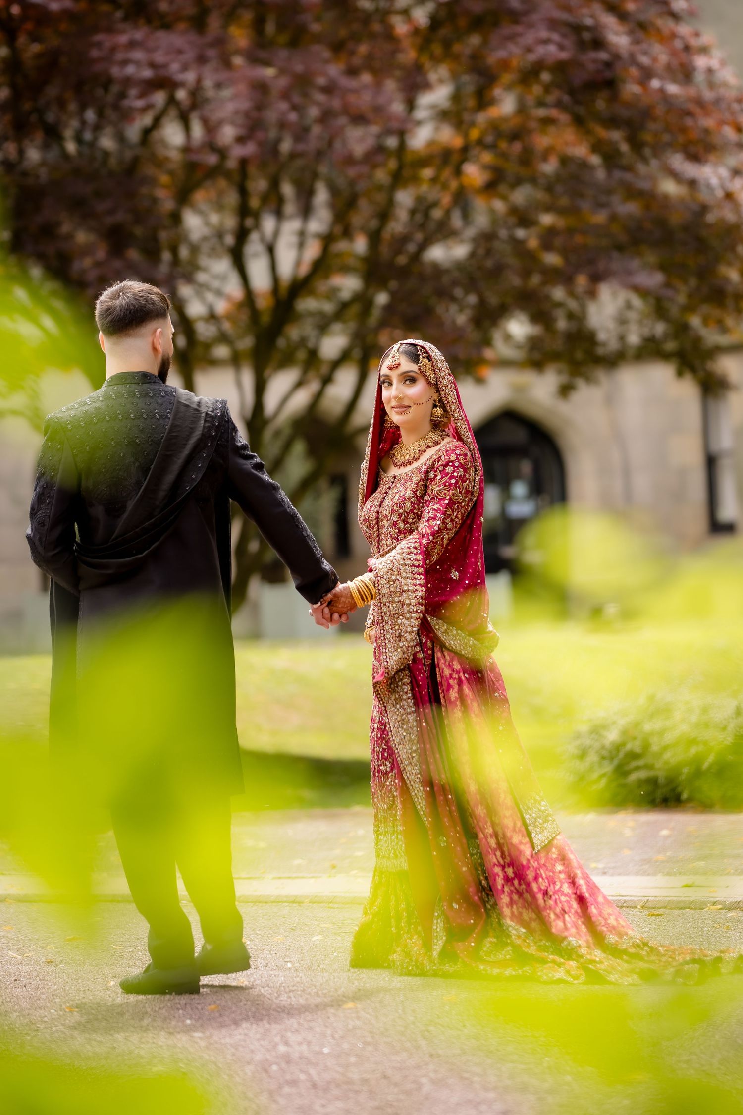 South Asian bride in red and gold lehenga holds groom's hand outdoors during traditional wedding photoshoot.