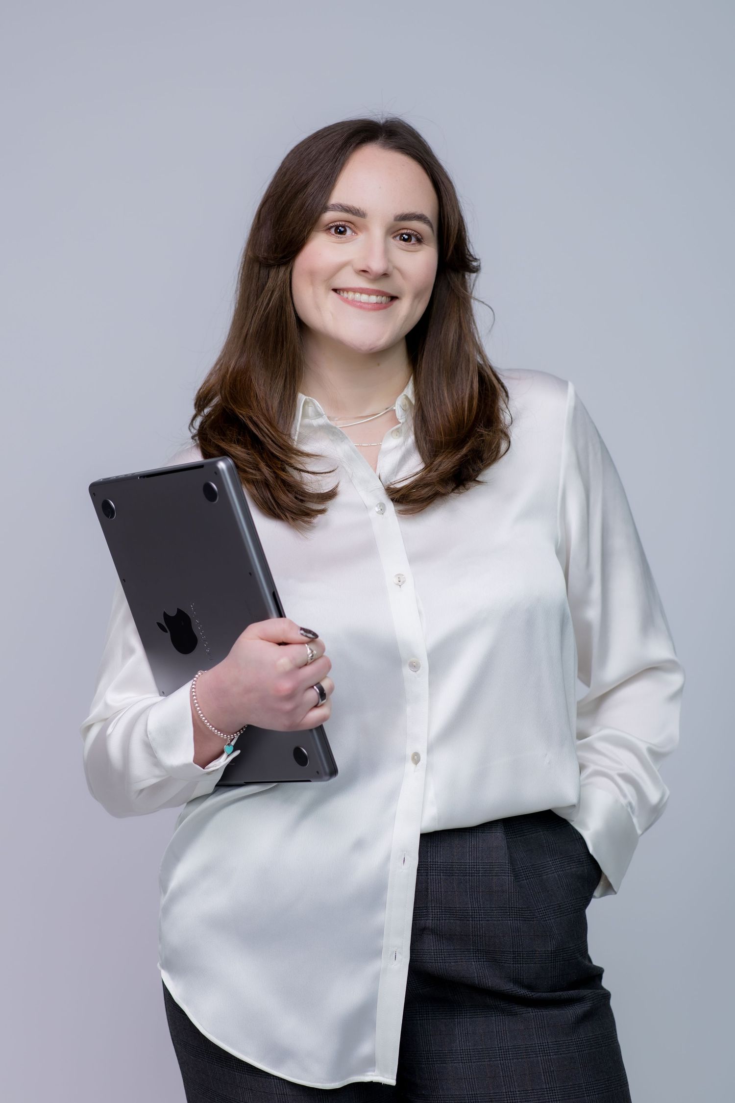 Smiling businesswoman in white blouse holding Apple laptop, standing confidently against gray background.