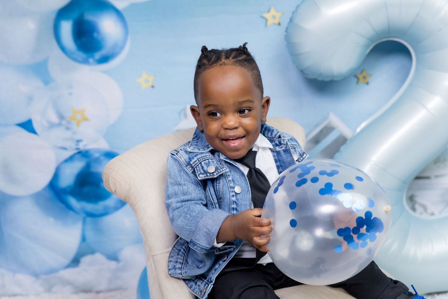 Smiling toddler in denim jacket holds balloon at blue-themed 2nd birthday party with star decorations.
