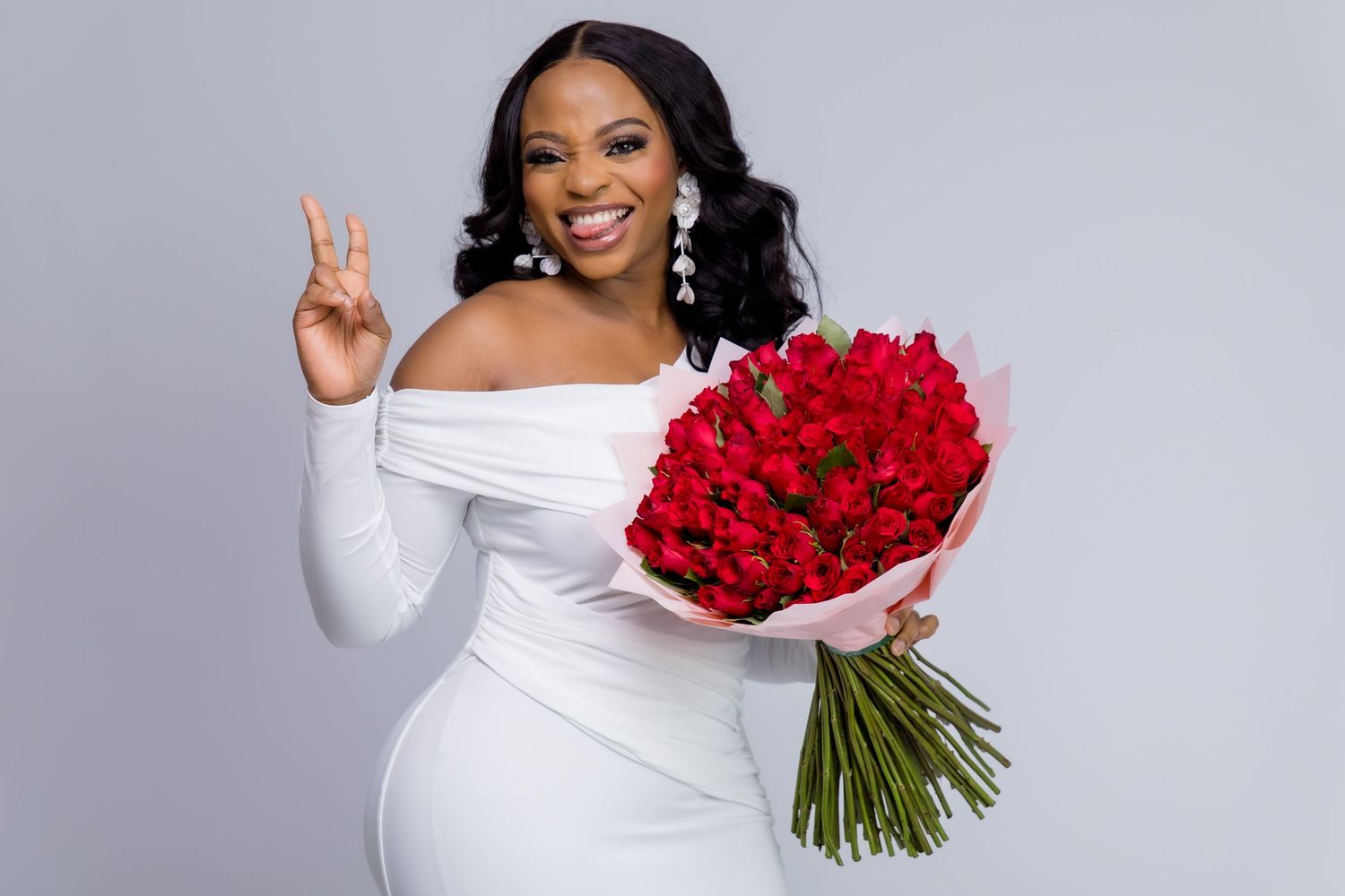 Woman in white off-shoulder dress holding red roses bouquet, making peace sign, smiling against gray background.