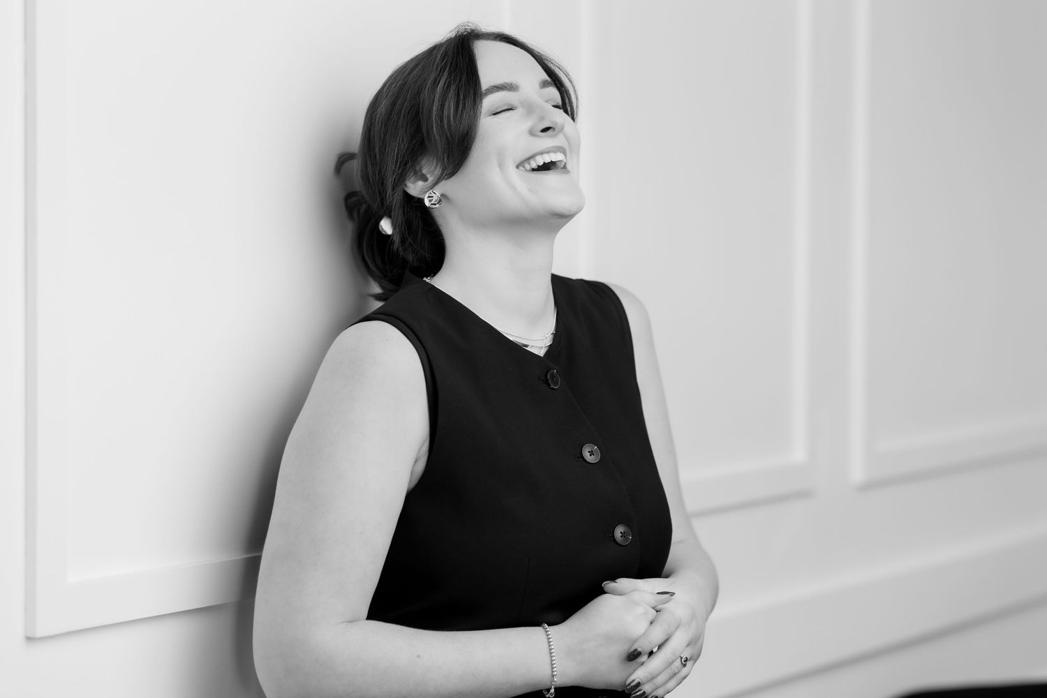 Black and white photo of a laughing woman in a sleeveless black dress leaning against a white wall.
